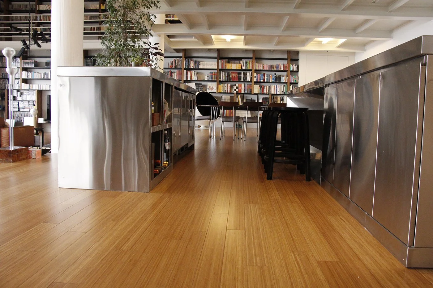 Interior of a modern library or bookstore with wooden flooring, metal countertops, black chairs, and shelves filled with books.