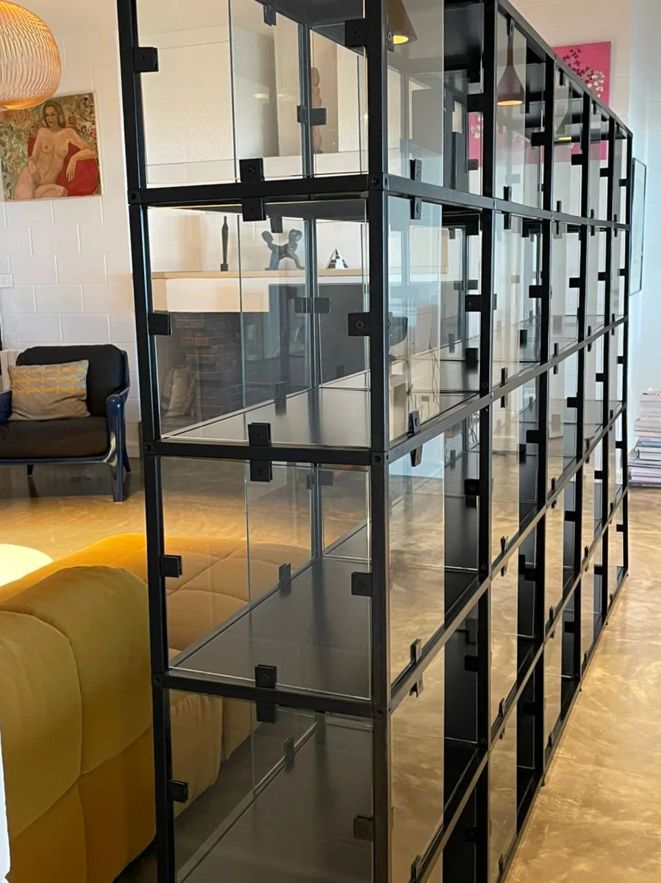 Image of a black metal and glass shelving unit in a living room with artwork, a fireplace, a sofa, and a yellow couch.