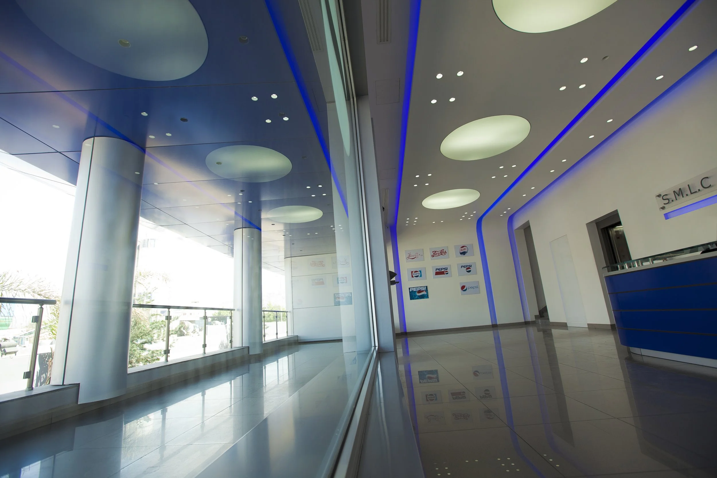 Modern interior space with shiny tiled floors, large windows, blue accent lighting, circular ceiling lights, and a white wall displaying Pepsi logos.