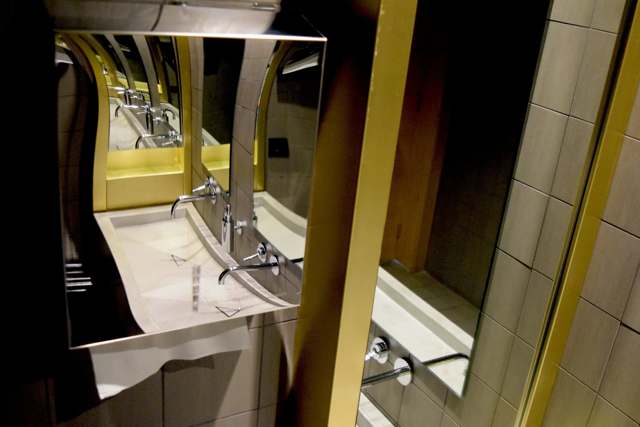 A bathroom with a yellow frame mirror above two sinks. The sinks have chrome faucets. An electrical outlet is visible near the mirror. The bathroom has beige tiles on the floor and walls, and a wooden element to the right.