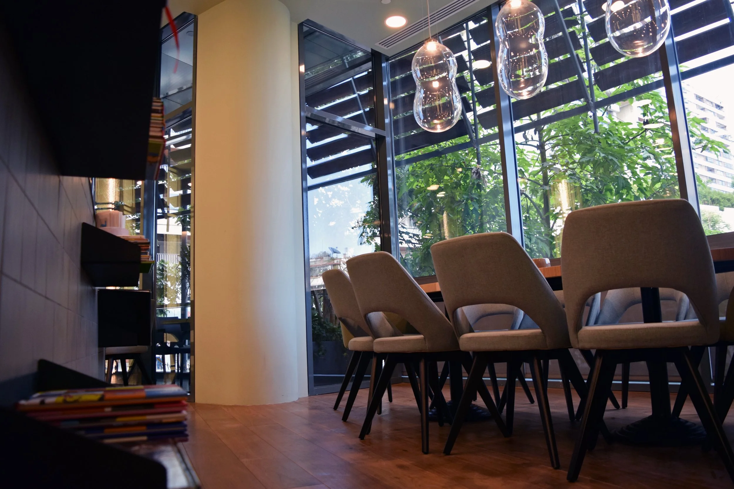 A modern indoor space with large floor-to-ceiling windows showing greenery outside. There are several beige chairs around a meeting or dining table. Hanging glass light fixtures are visible from the ceiling, and shelves with books are seen on the lef