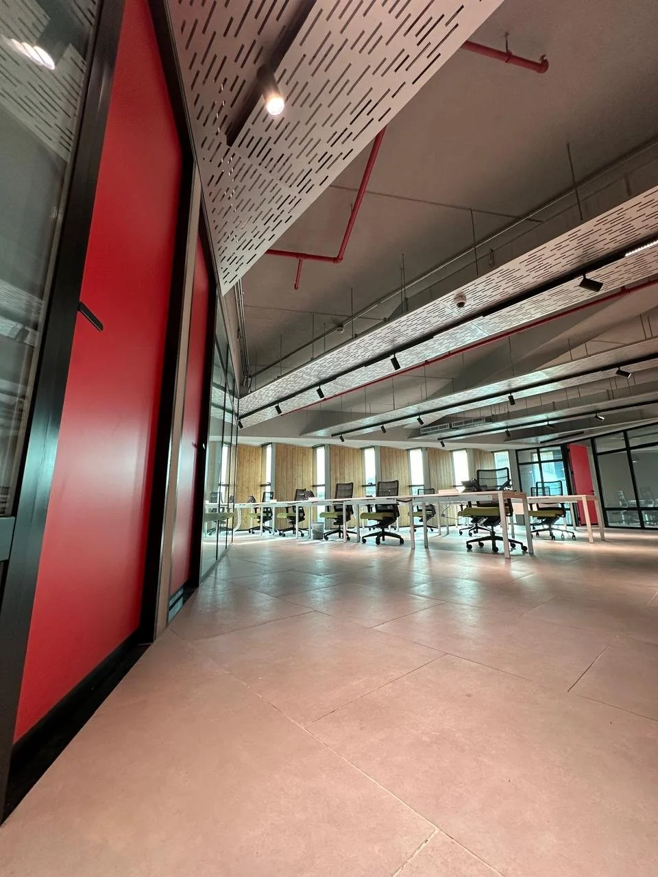 An empty modern conference room with office chairs and long tables, large windows, and a ceiling with exposed red piping and distinctive ceiling panels.