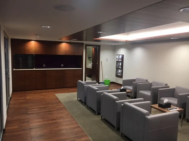 Empty lounge area with nine gray armchairs arranged in three rows, a small rectangular wooden table with a tissue box, a green cleaning bucket, and a wall-mounted photo collage, with a wooden panel wall and a retractable glass door.
