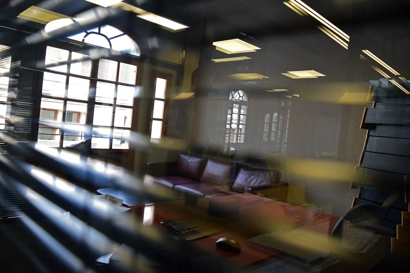 Office conference room seen through window blinds, with windows, a leather couch, a table with a keyboard and mouse, and shelves.