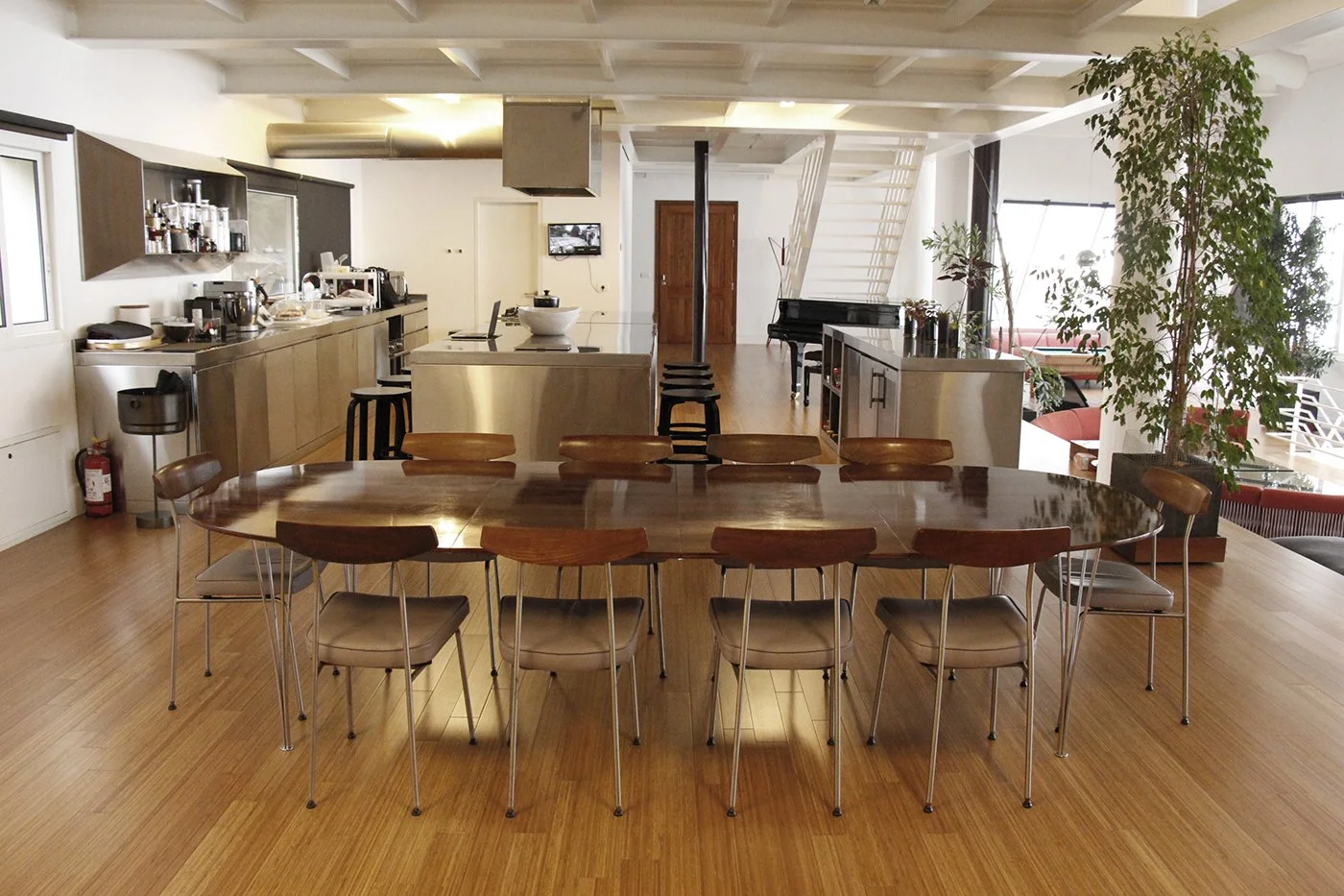 Spacious open-concept kitchen and dining area with wooden flooring, a large oval wooden dining table, modern kitchen with island, black chairs, potted plants, a piano, and natural light.