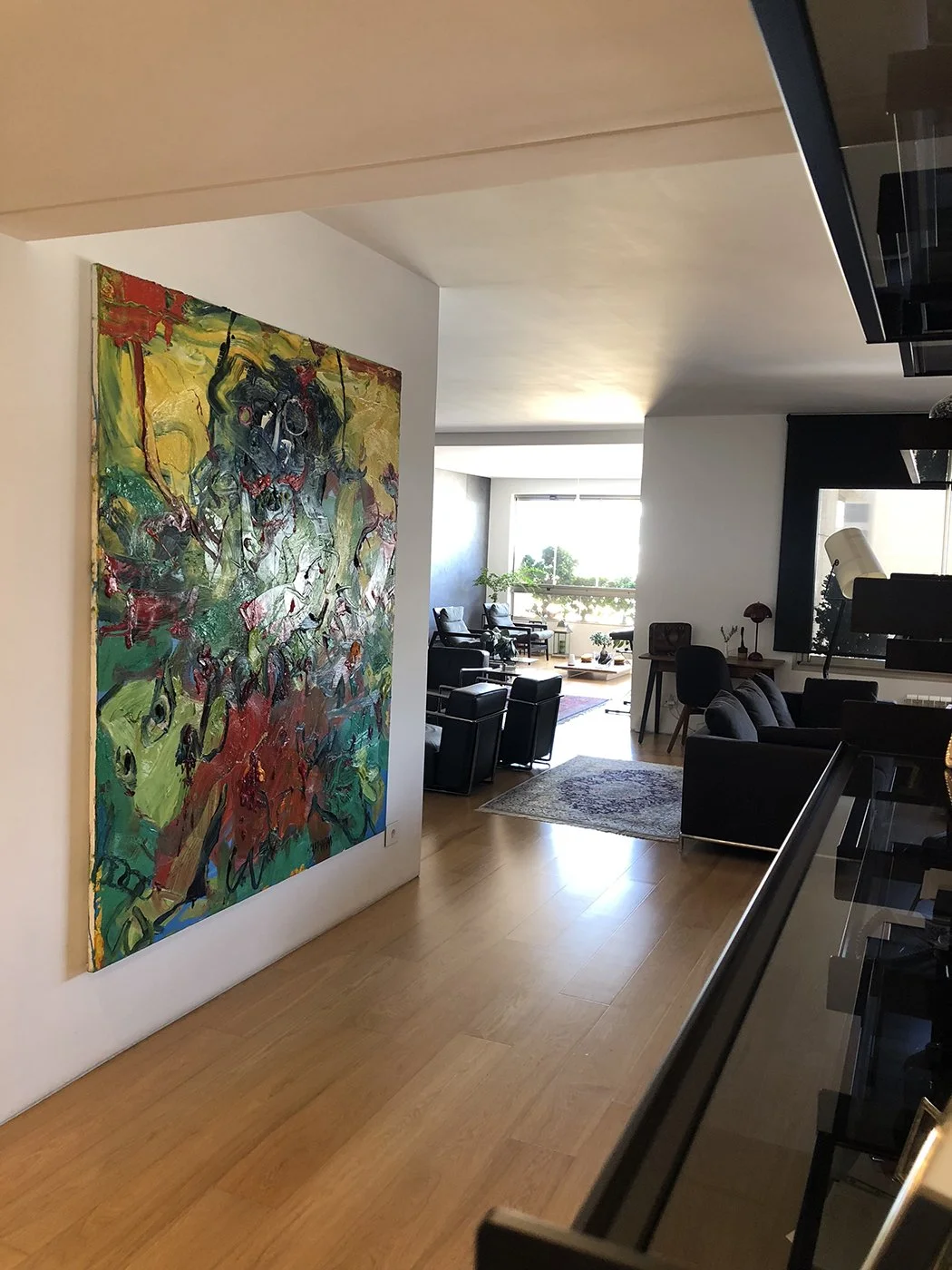 Modern living room with large colorful abstract painting on white wall, black armchairs, sofa, and large window with plants outside.