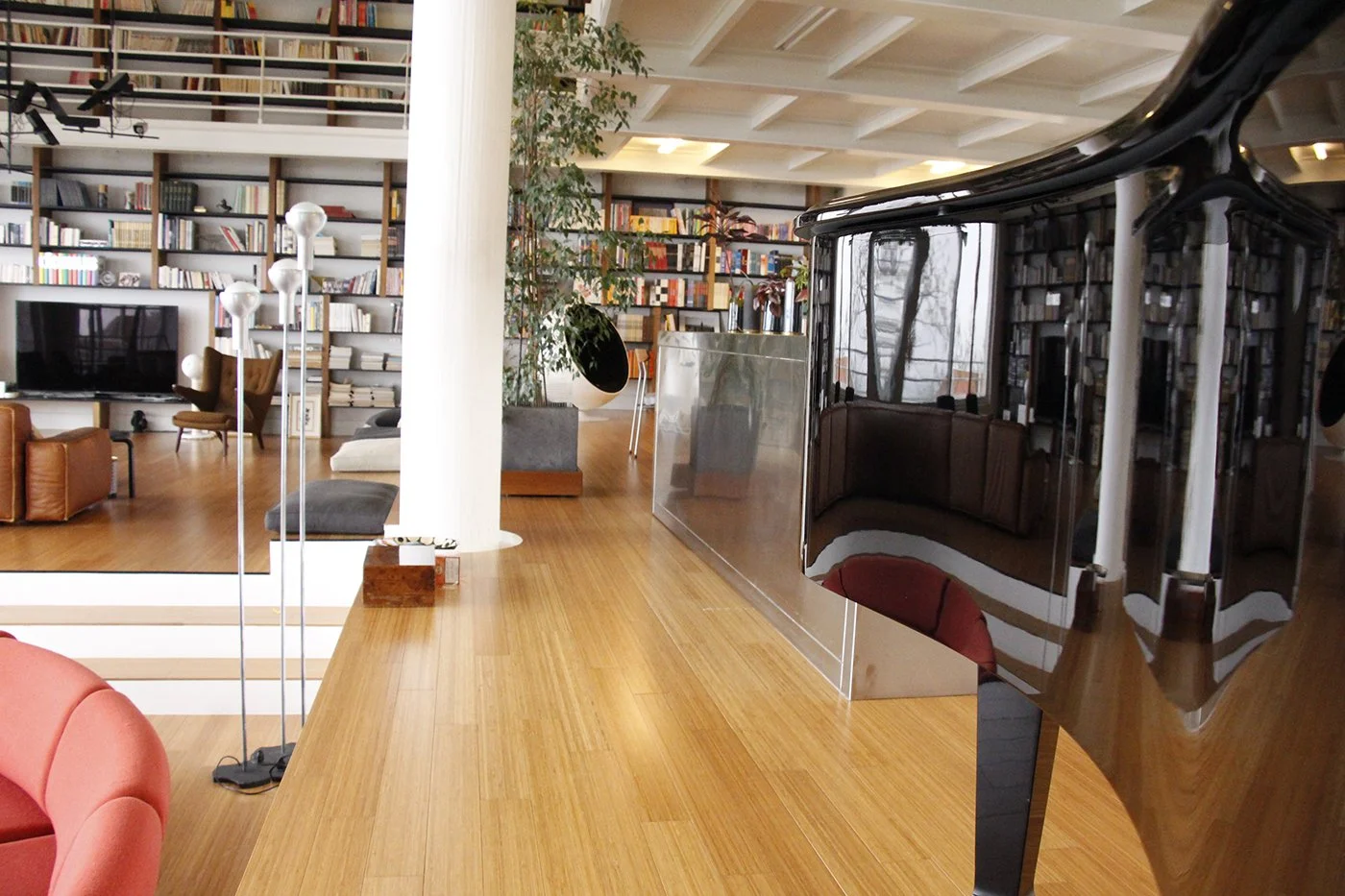 Interior of a modern living room with a grand piano, wooden flooring, bookshelves, plants, and various seating areas.