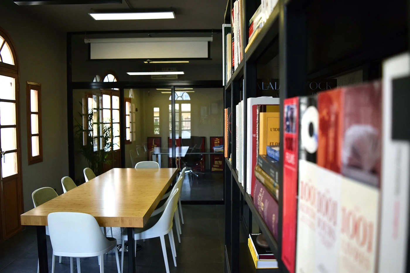 A library conference room with a long wooden table and white chairs, windows with wooden frames, and bookshelves in the foreground.