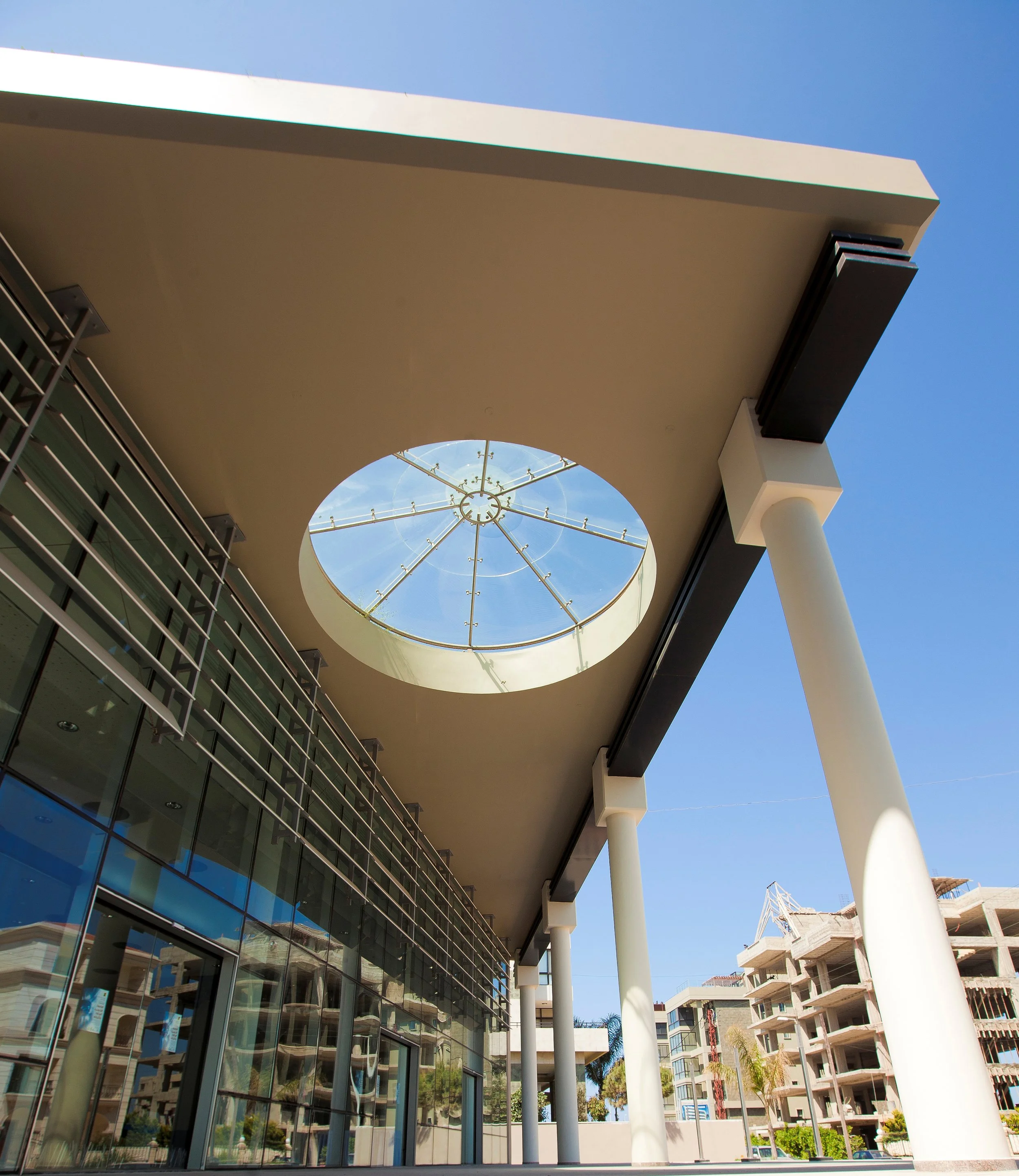 Modern building with large glass windows, white columns, and a circular glass skylight in a sunny urban area with high-rise buildings in the background.
