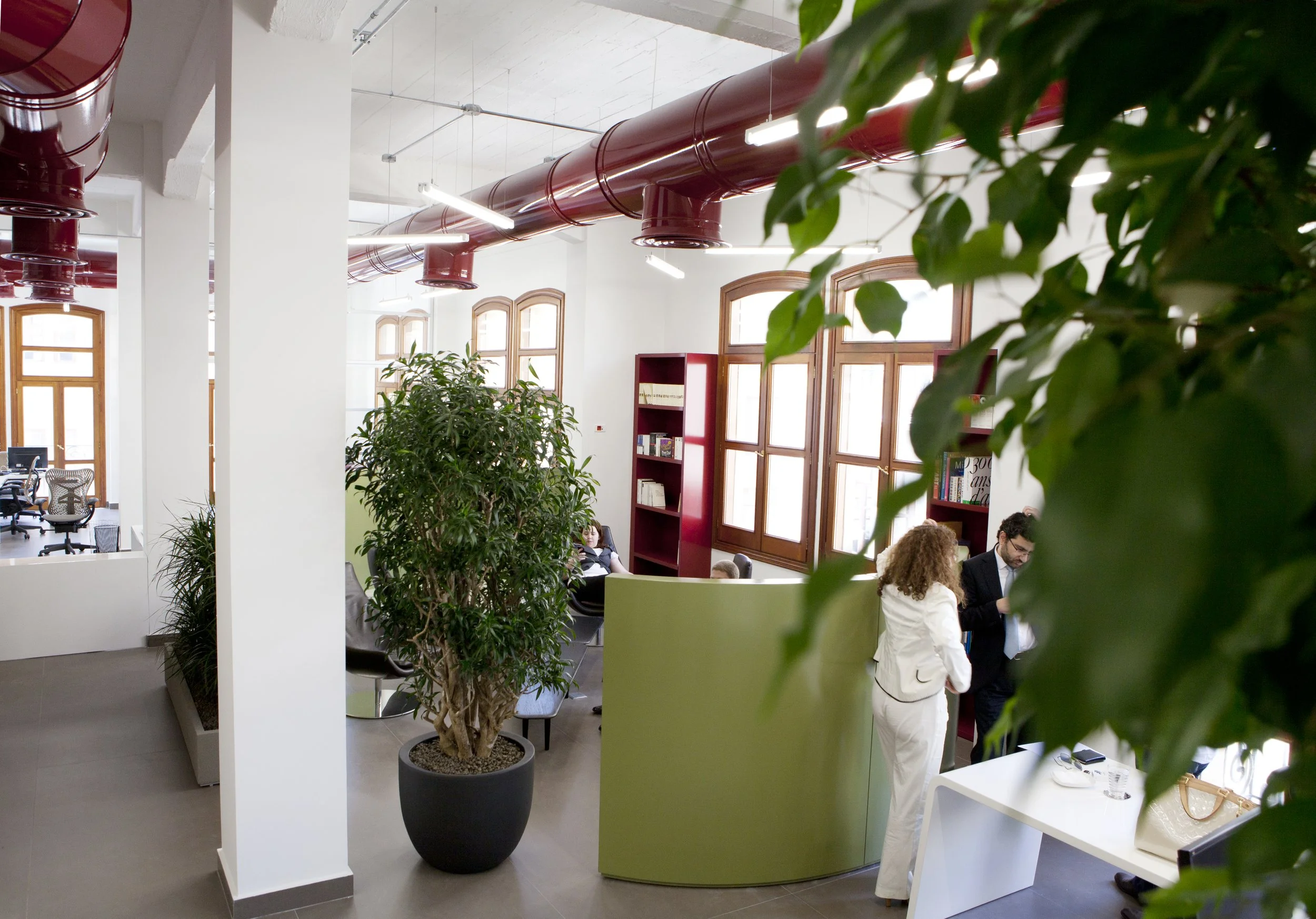 Modern office space with large potted plants, red ductwork along the ceiling, and several people engaged in conversation near a green partition.