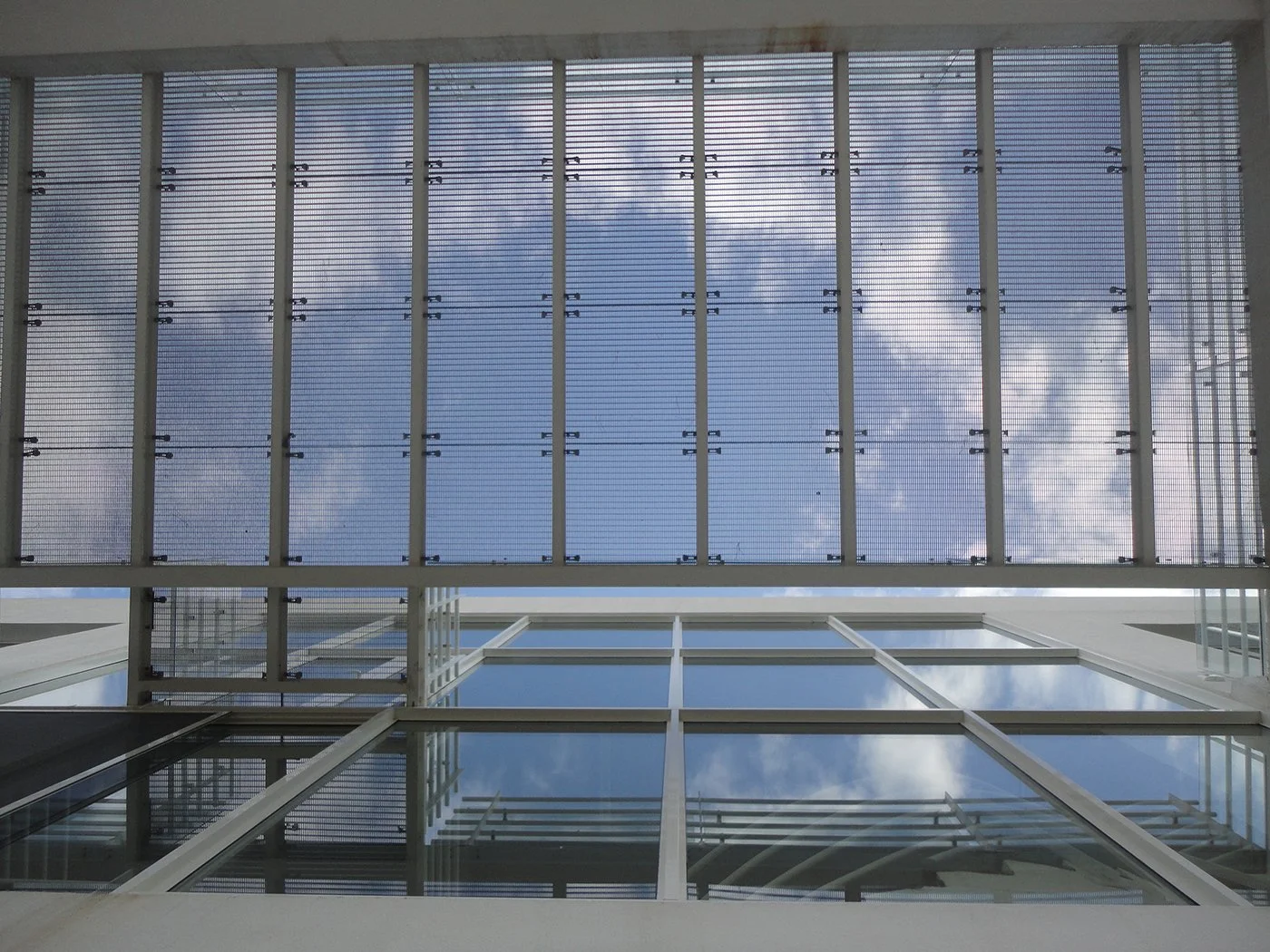 Looking up at a building's exterior, showing glass windows on the lower part and a metal grid or sunshade on the upper part with a blue sky and some clouds visible.