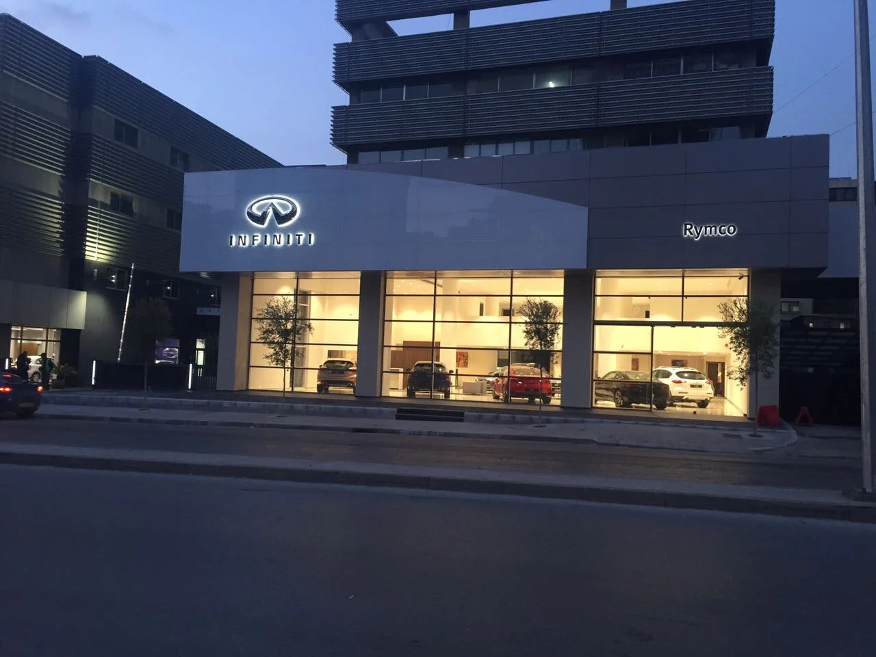 A modern car dealership building with illuminated Infiniti logo and cars inside visible through large glass windows, evening setting with a nearby street.