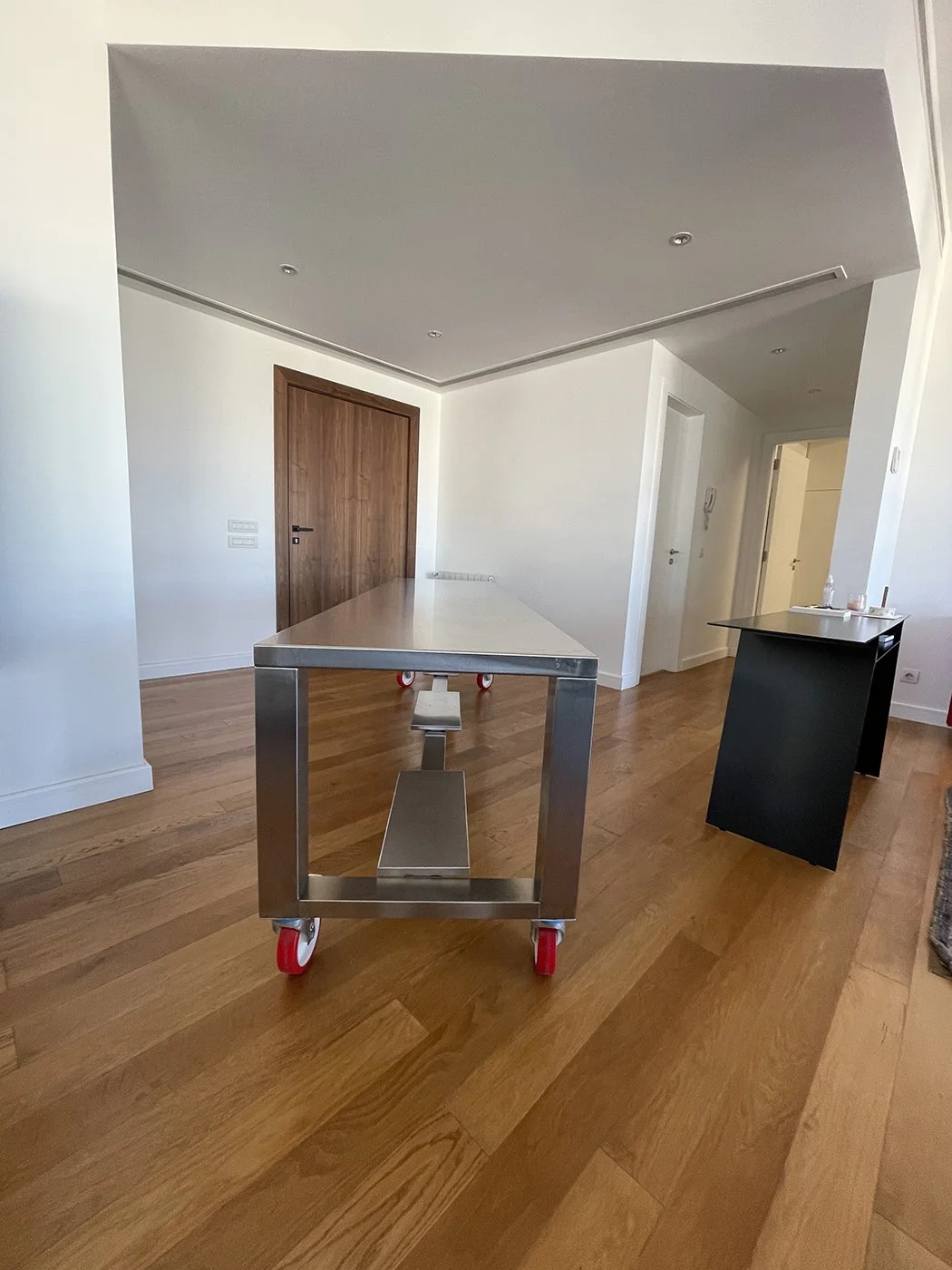A modern kitchen or dining area with a wooden door, white walls, and wooden flooring. There is a metal cart with red wheels in the center and a black cabinet or table on the right side.