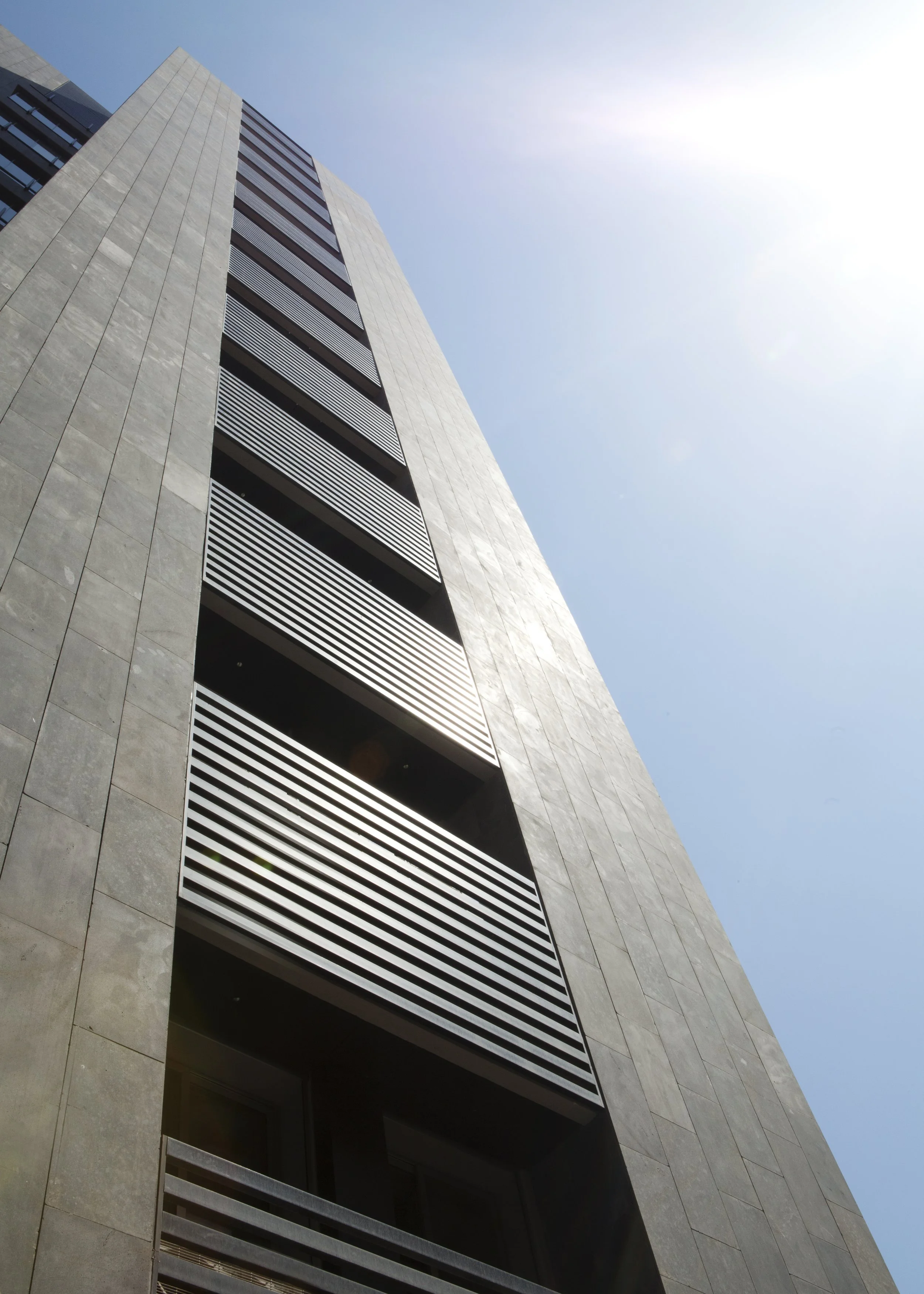 Low-angle view of a modern high-rise building with horizontal metal louvers on the balconies, against a clear blue sky with the sun shining brightly.