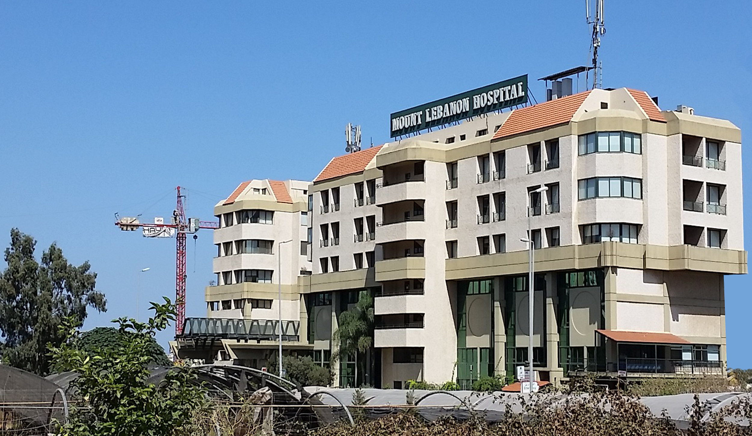 A multi-story building with a sign that reads 'Mount Lebanon Hospital' on the roof, under a blue sky. There are construction cranes and trees in front of the building.