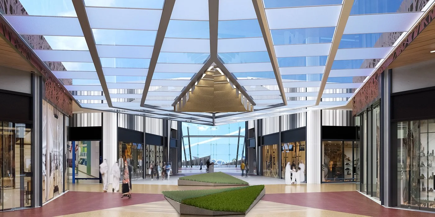 Modern shopping mall with a glass ceiling, storefronts on either side, and people walking through the open center area with landscaped grass sections.