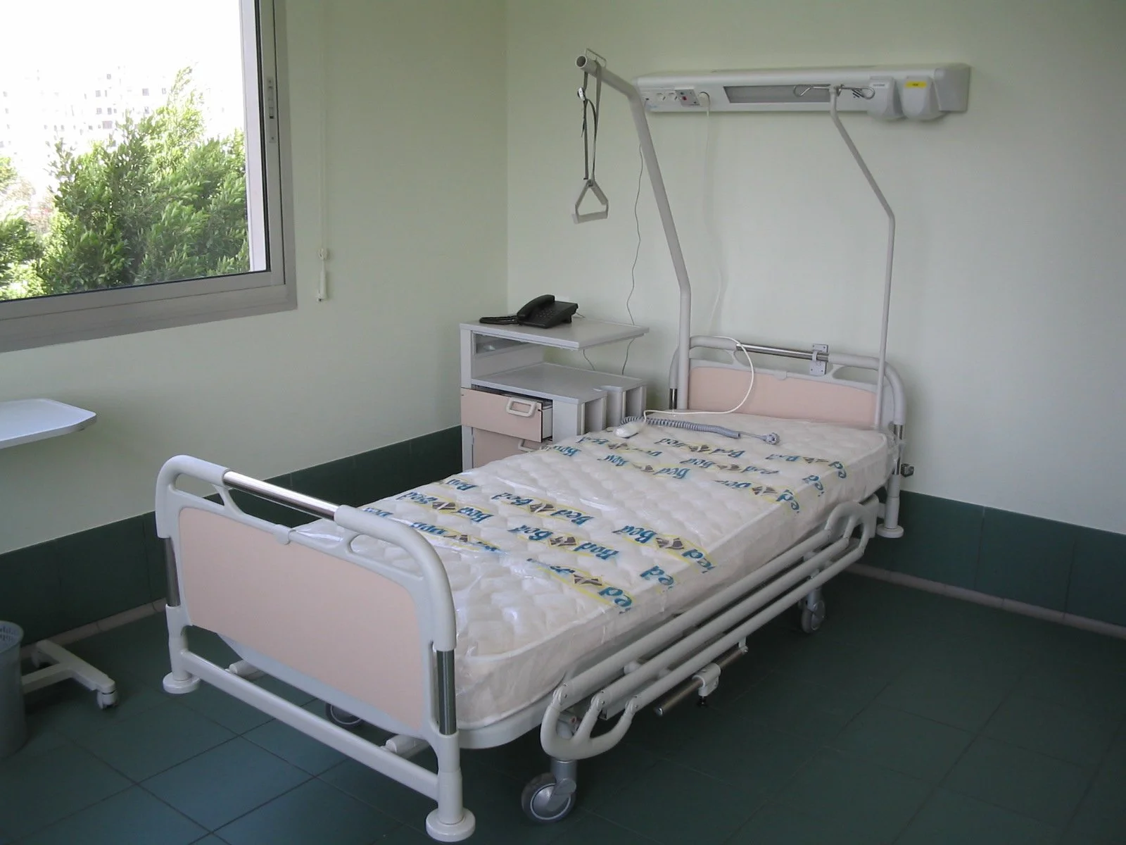 Hospital room with an empty bed, window showing trees outside, and medical equipment mounted on the wall.