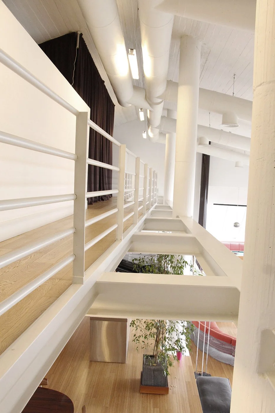 Interior view of a modern building with white beams, pipes on the ceiling, a wooden floor, a white railing, potted plants, and couches in a lounge area.