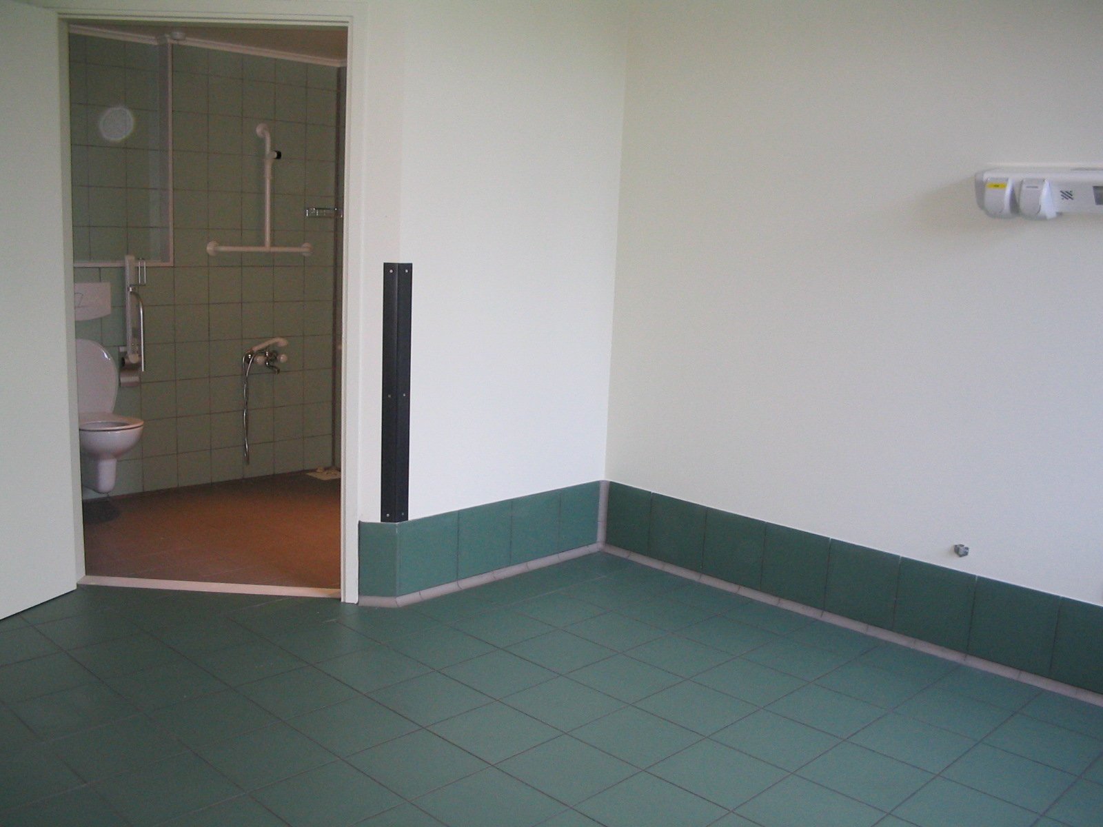 Empty room with green tiled floor, white walls, and a doorway leading to a wheelchair-accessible bathroom with green tile walls, a toilet, and grab bars.