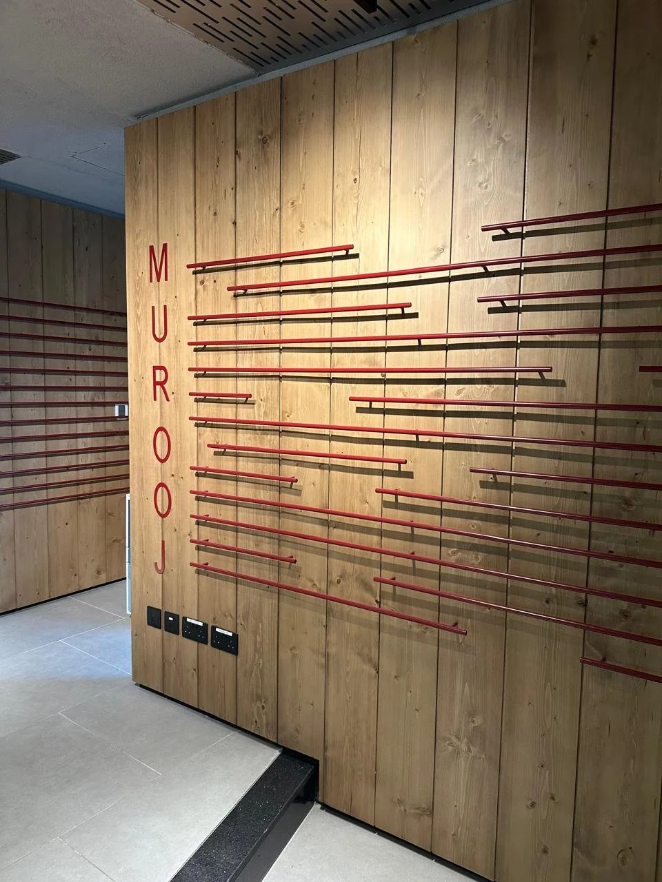 Wood-paneled wall with red and black metal rods and the word 'MURJOJ' written vertically in red letters.