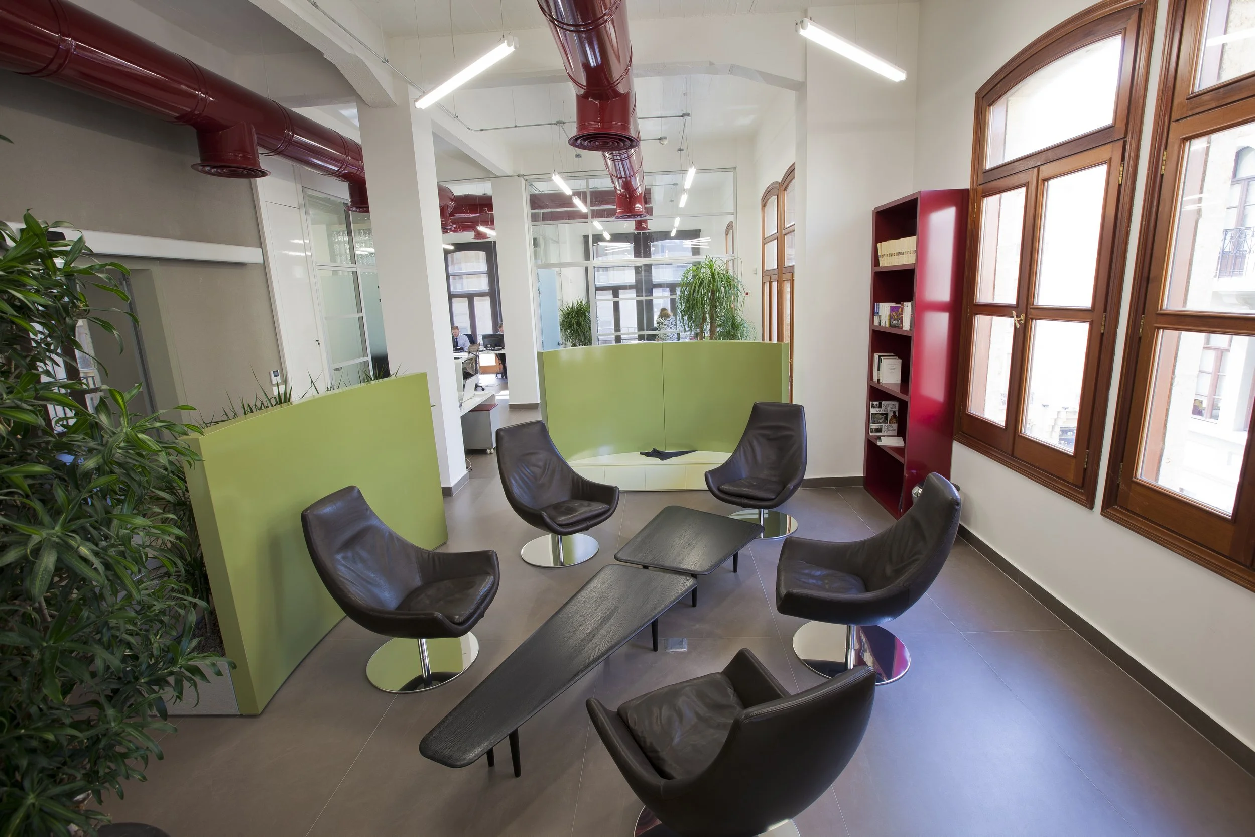 A modern office lounge area with six black leather chairs and a small rectangular table in the center. Green and red partition walls, large wooden framed windows, and indoor plants are visible. In the background, there are employees working at desks.