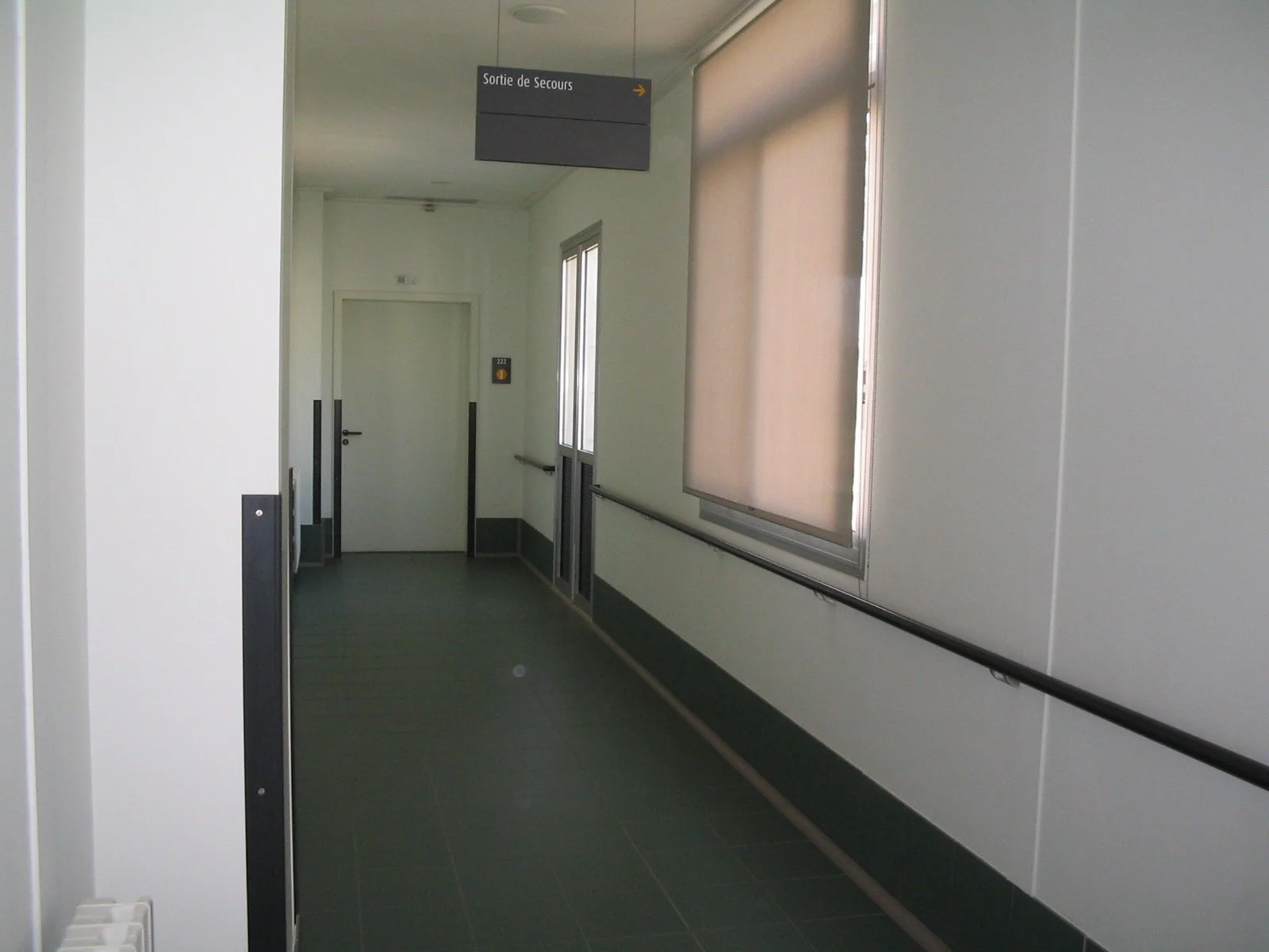Hospital corridor with gray floor, white walls, black handrails, a window with closed blind, and a sign pointing to the emergency exit.