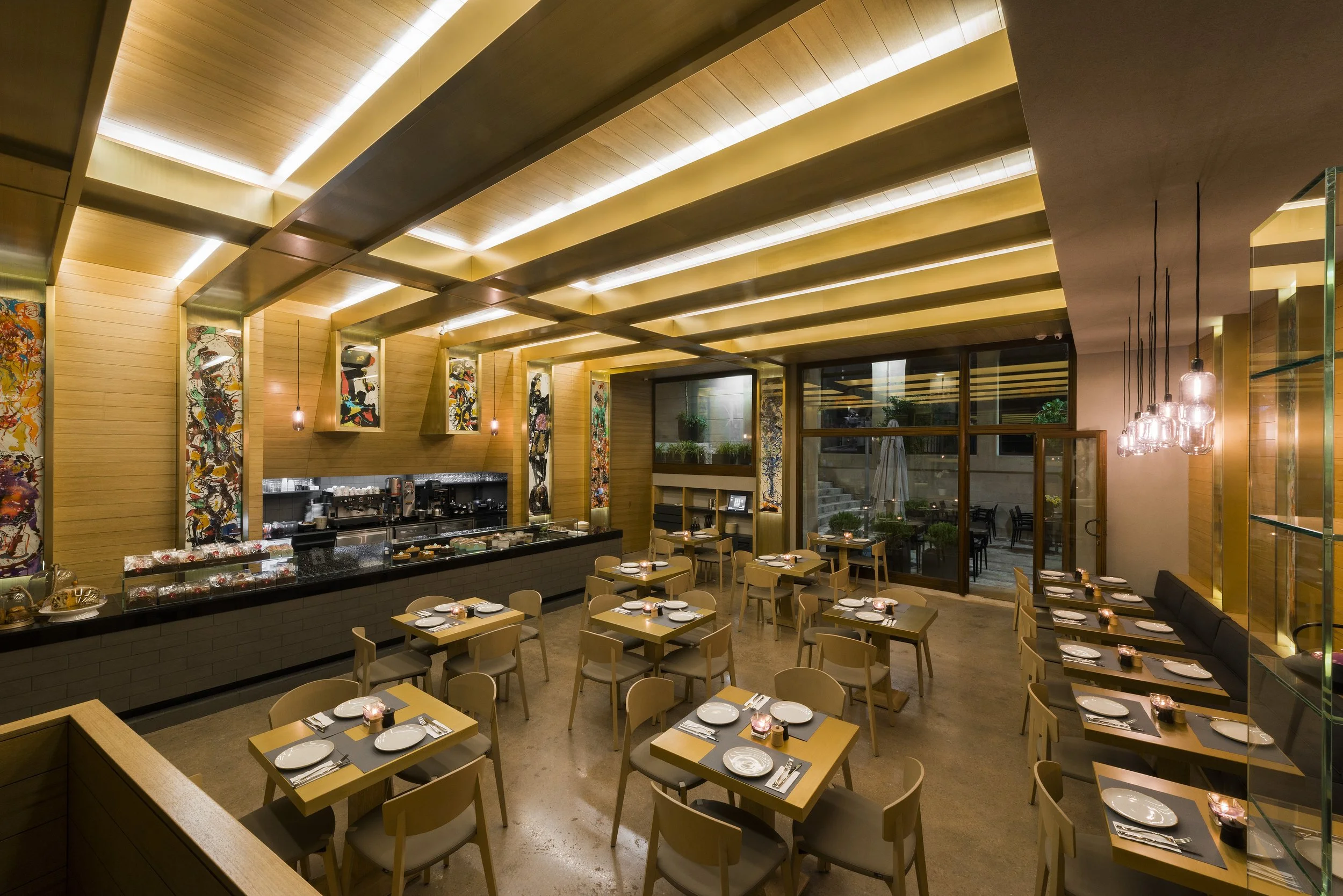 Interior of a modern restaurant with wooden walls and ceiling, decorated with colorful abstract artwork, tables set with plates and candles, and a coffee station in the background.