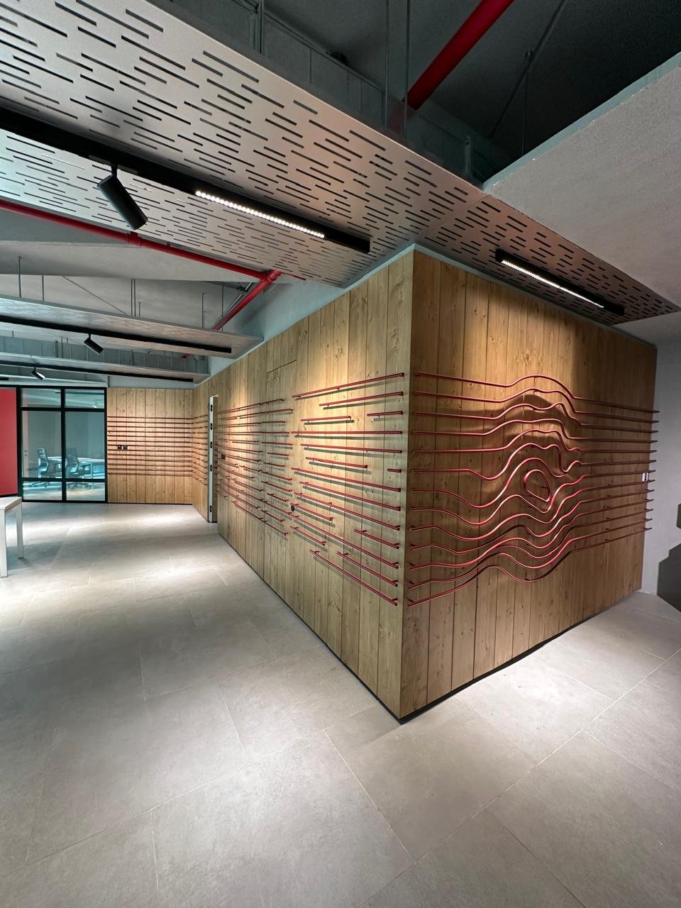 Interior of a modern building with a wooden corner wall decorated with red metal lines arranged in abstract wave patterns. The ceiling has a perforated wooden panel with integrated lighting and black track lights. Glass doors are visible in the background.