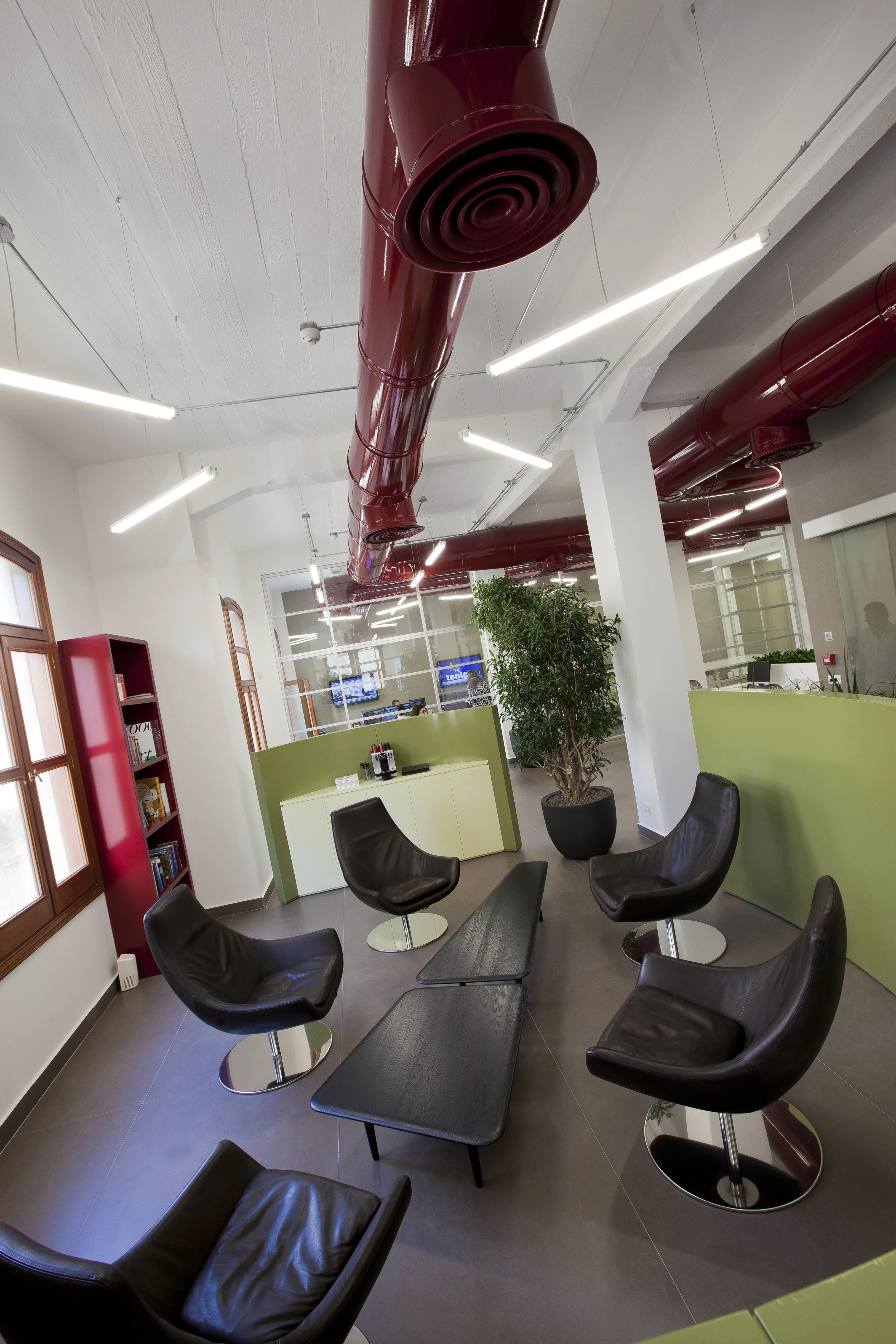 Modern office lounge with black leather chairs, a black coffee table, green accents, a large plant, bookshelves, and red exposed ductwork on the ceiling.