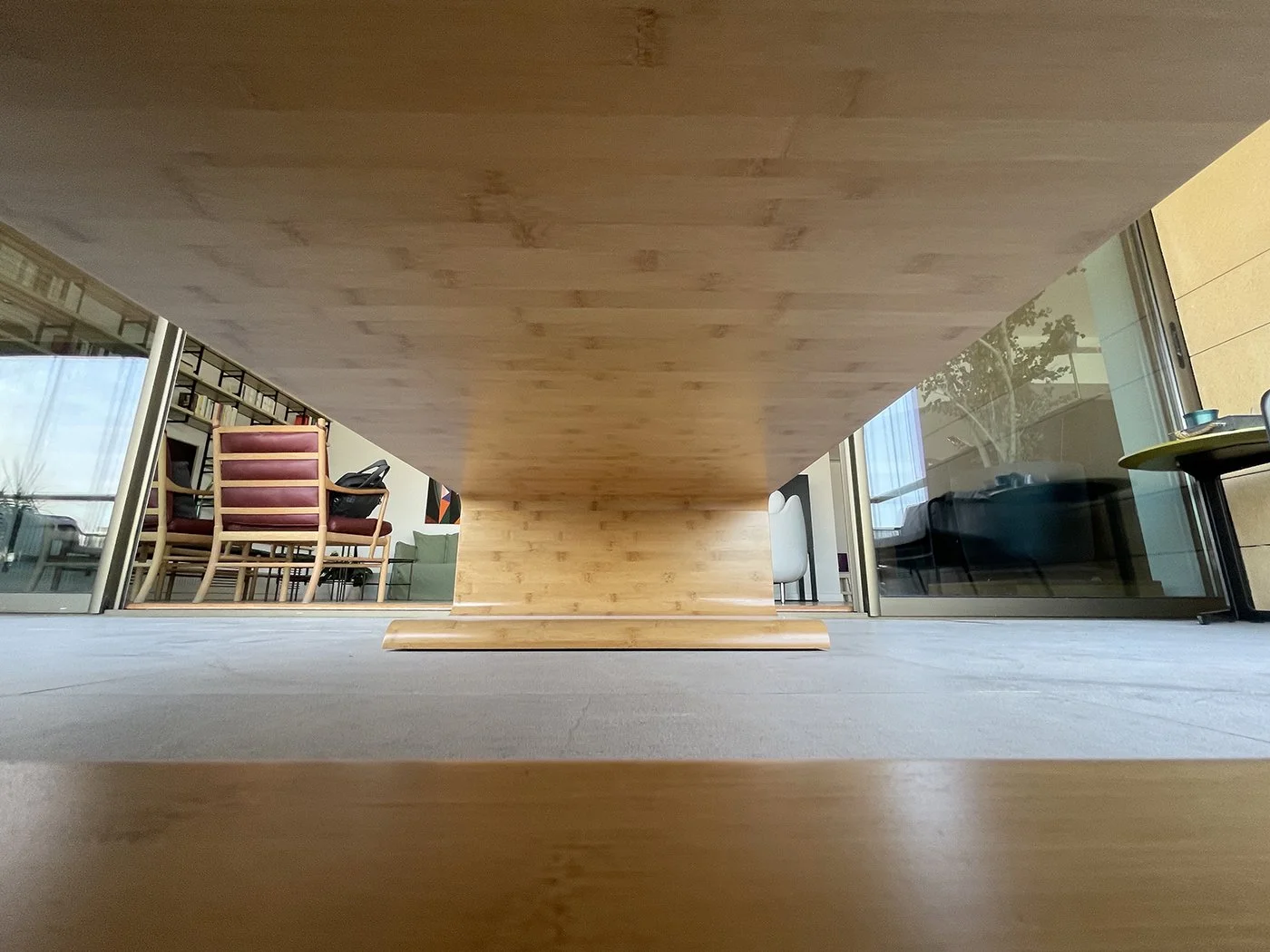 Inside a modern room with a wooden table seen from below, with chairs and a black desk phone on a chair to the left, and glass sliding doors revealing an outdoor patio and a sitting area with a green couch and window with curtains.