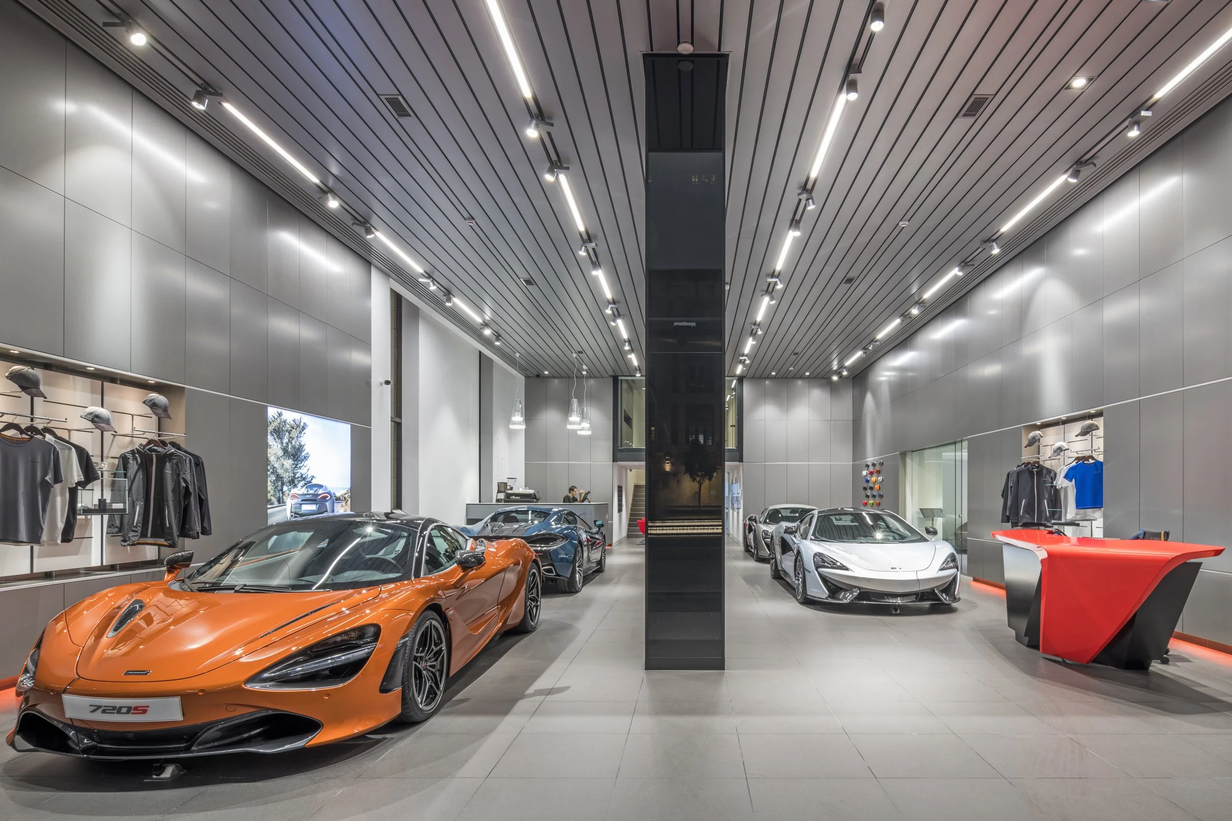 Inside a modern car showroom with luxury sports cars, including a prominent orange McLaren 720S in the foreground and other high-end cars displayed behind, with sleek lighting and clothing merchandise on the walls.