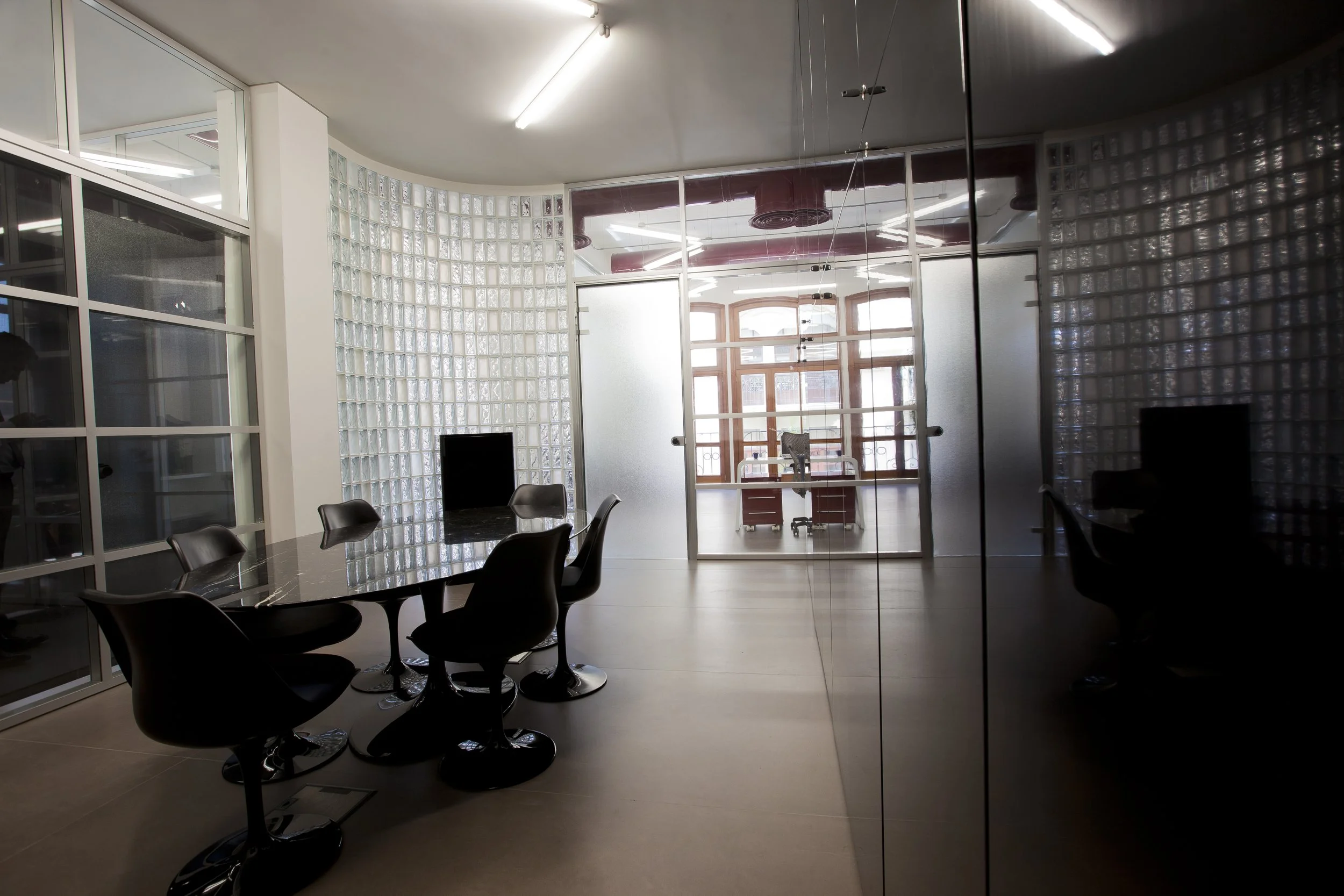 Modern conference room with black chairs around a dark marble table, glass block walls, large windows with wooden frames, and natural light.