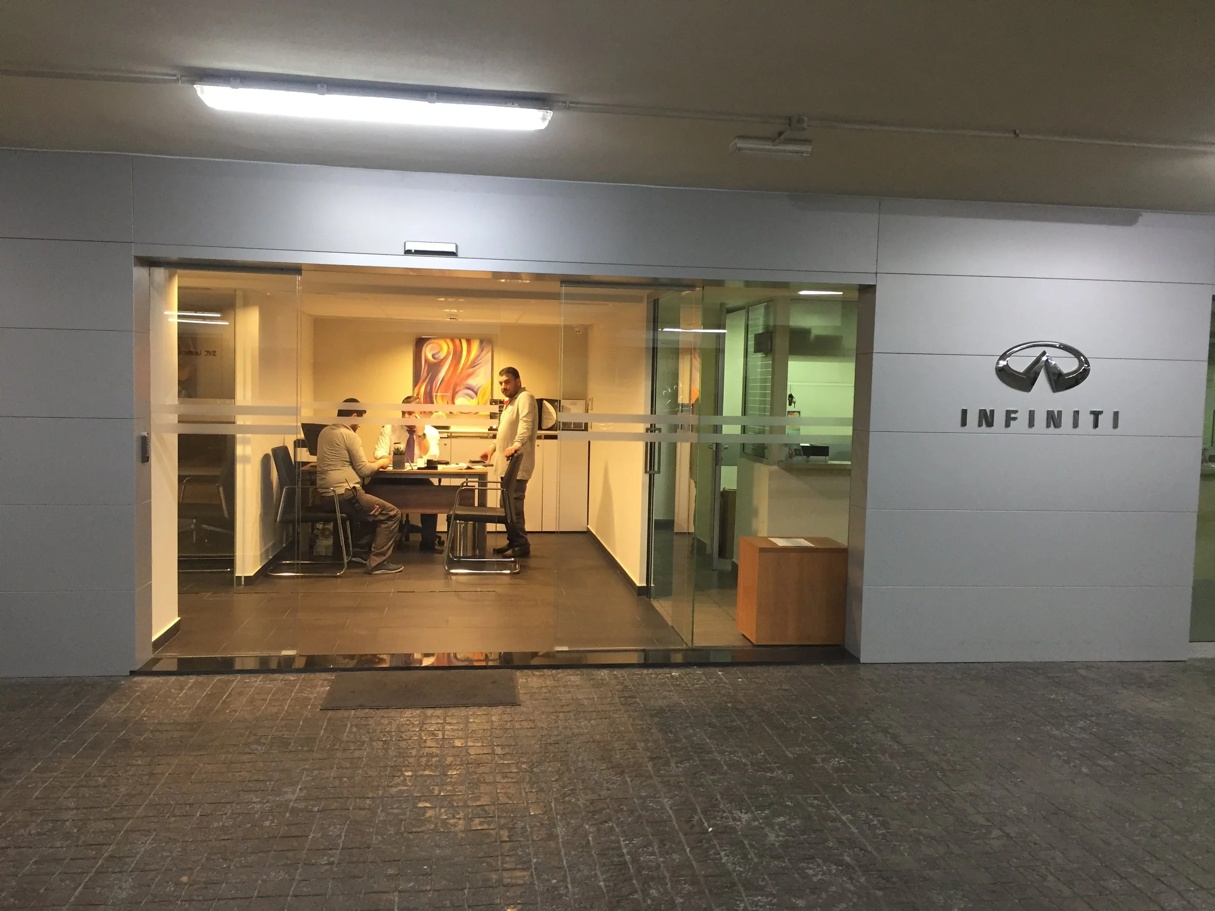 View of an Infiniti car dealership entrance with indoor office visible through glass doors, showing people working and a colorful abstract painting on the wall.