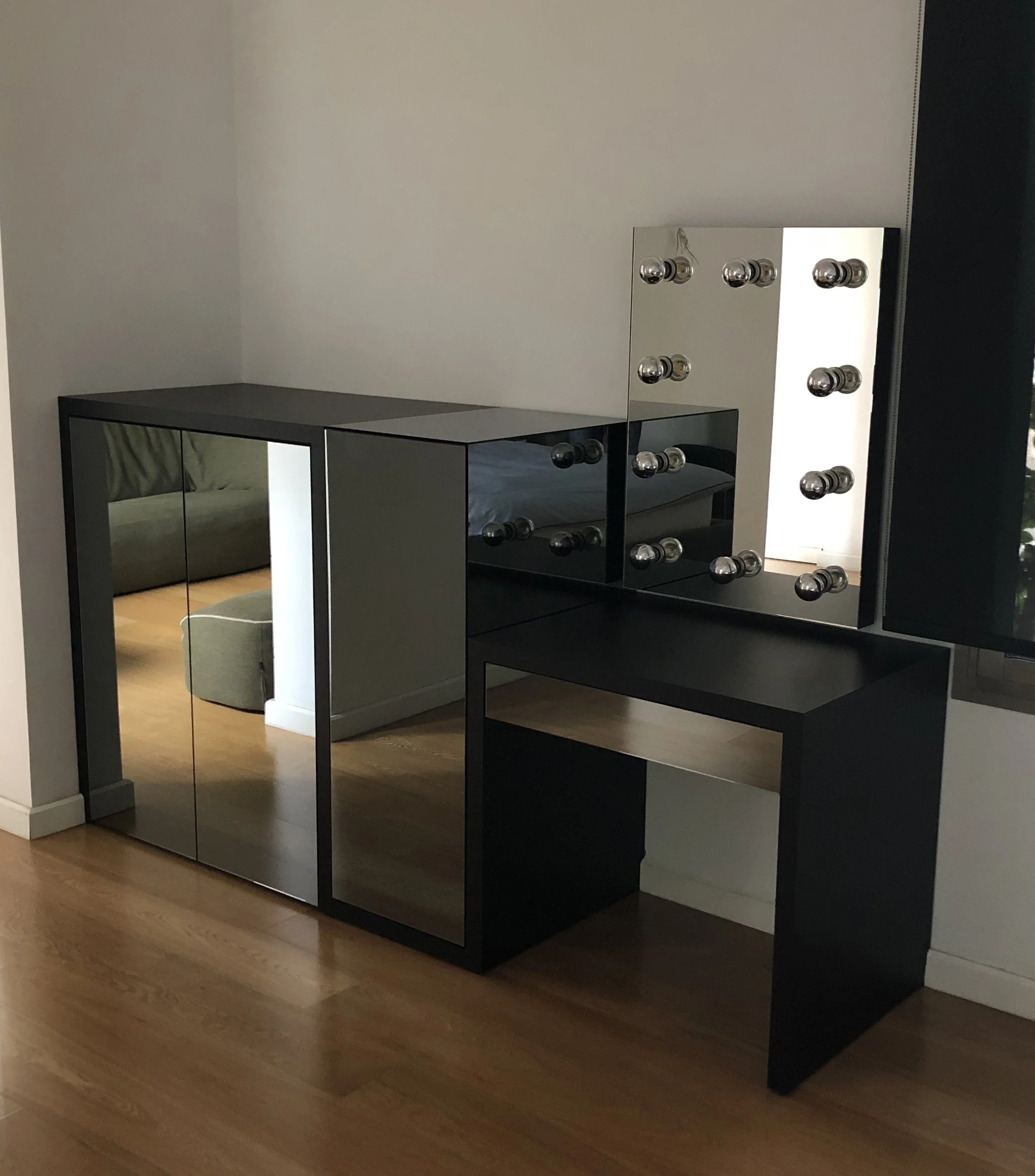A black makeup vanity with mirrored doors and a large mirror with round bulbs around the edges, placed in a room with wooden flooring and a white wall.