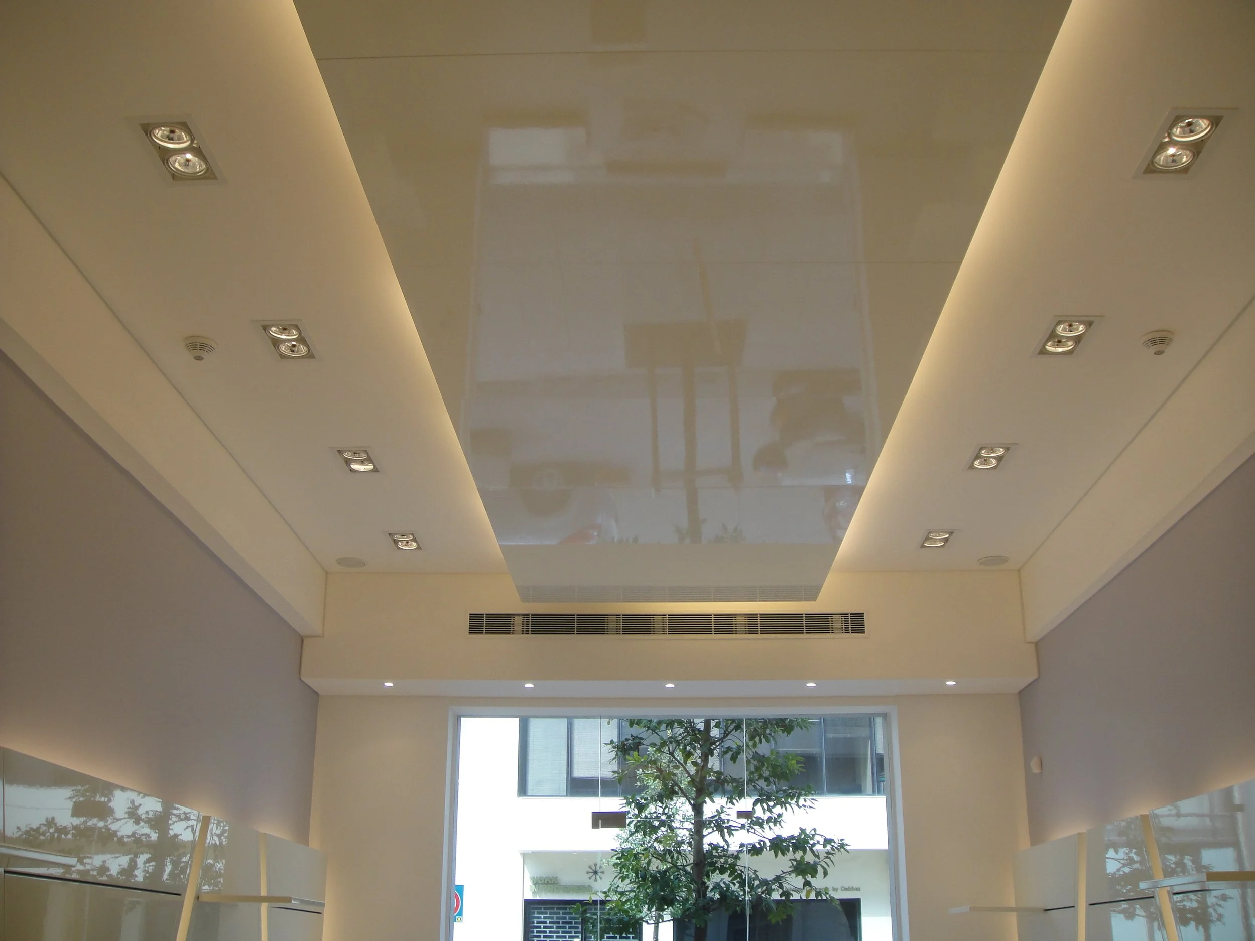 Interior view of a high-ceiling room with a large window at the far end, showing a tree outside. The ceiling has a shiny, reflective surface with embedded lights and a vent, and the walls are painted a light color.