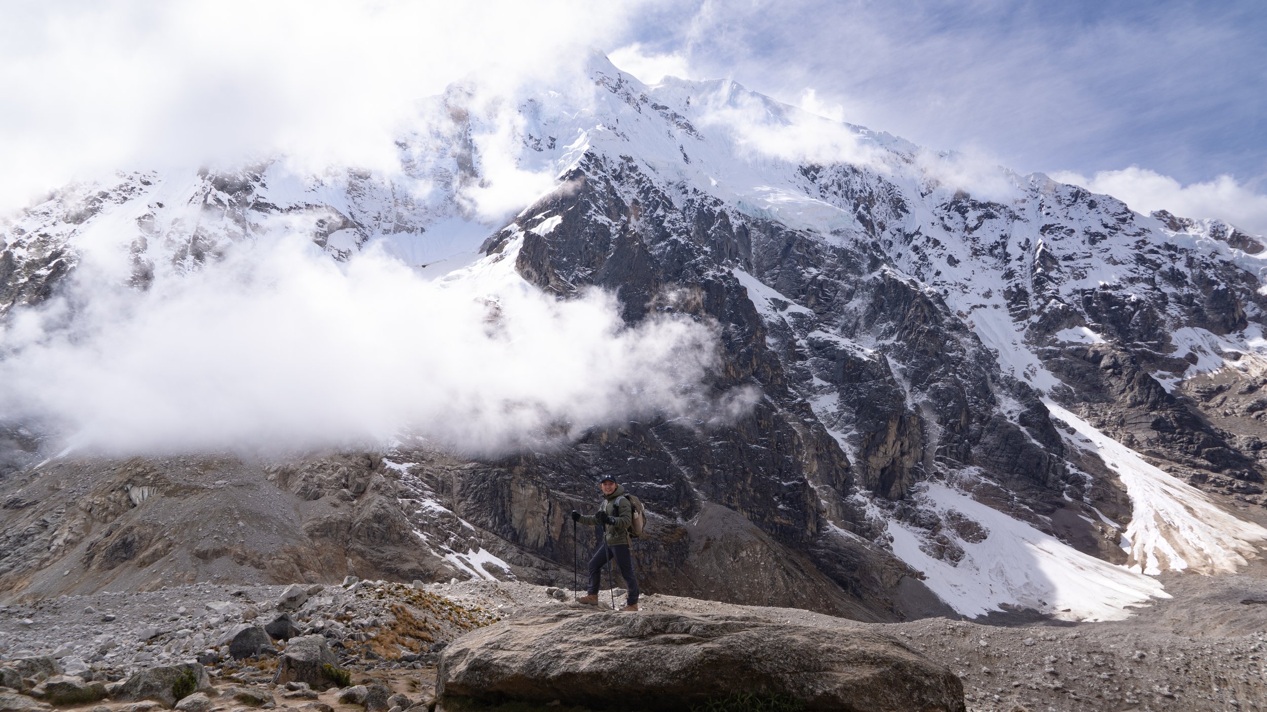 A man with hiking gear and poles standing on rocky terrain in front of a snowy mountain with clouds around its peak.
