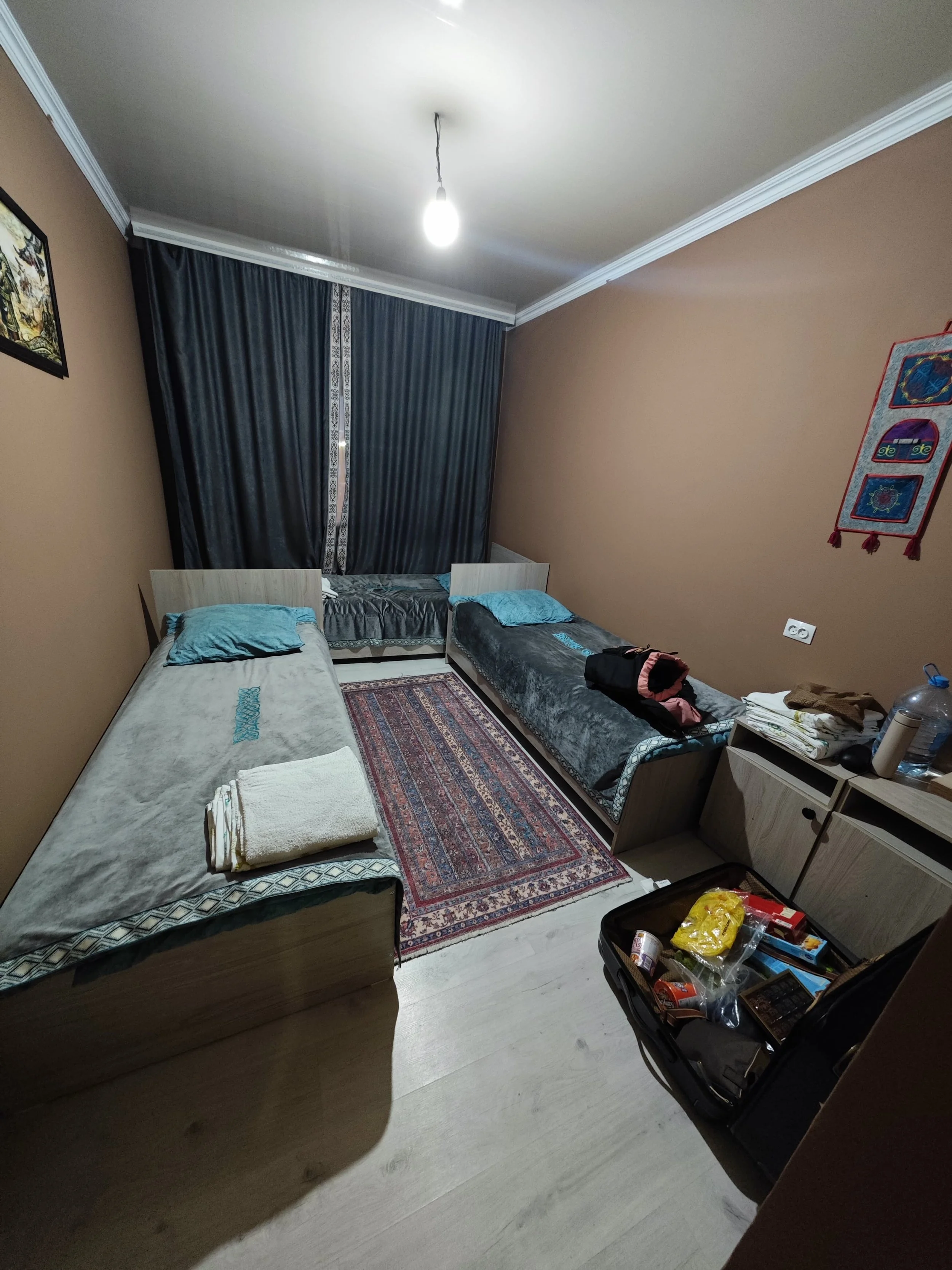 A bedroom with beige walls and white ceiling, featuring two single beds with blue pillows, a colorful rug, a nightstand with various items, and dark curtains covering the window. There is a lantern-style ceiling light turned on and a box of snacks on the floor.
