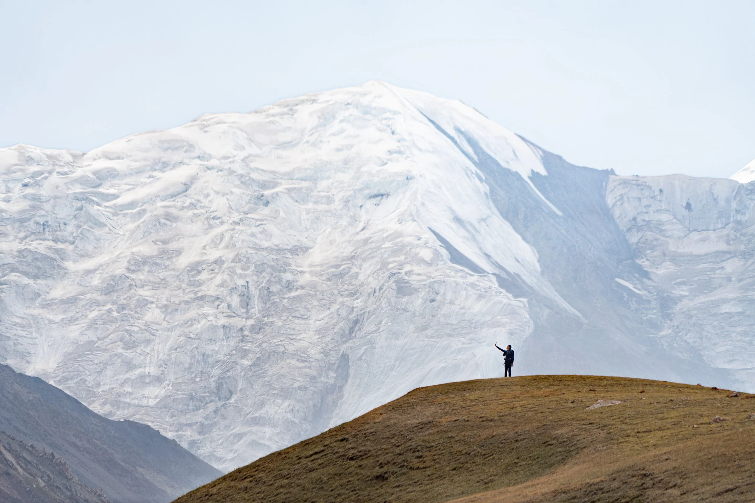 Person standing on a grassy hill taking a selfie with a snow-capped mountain in the background.