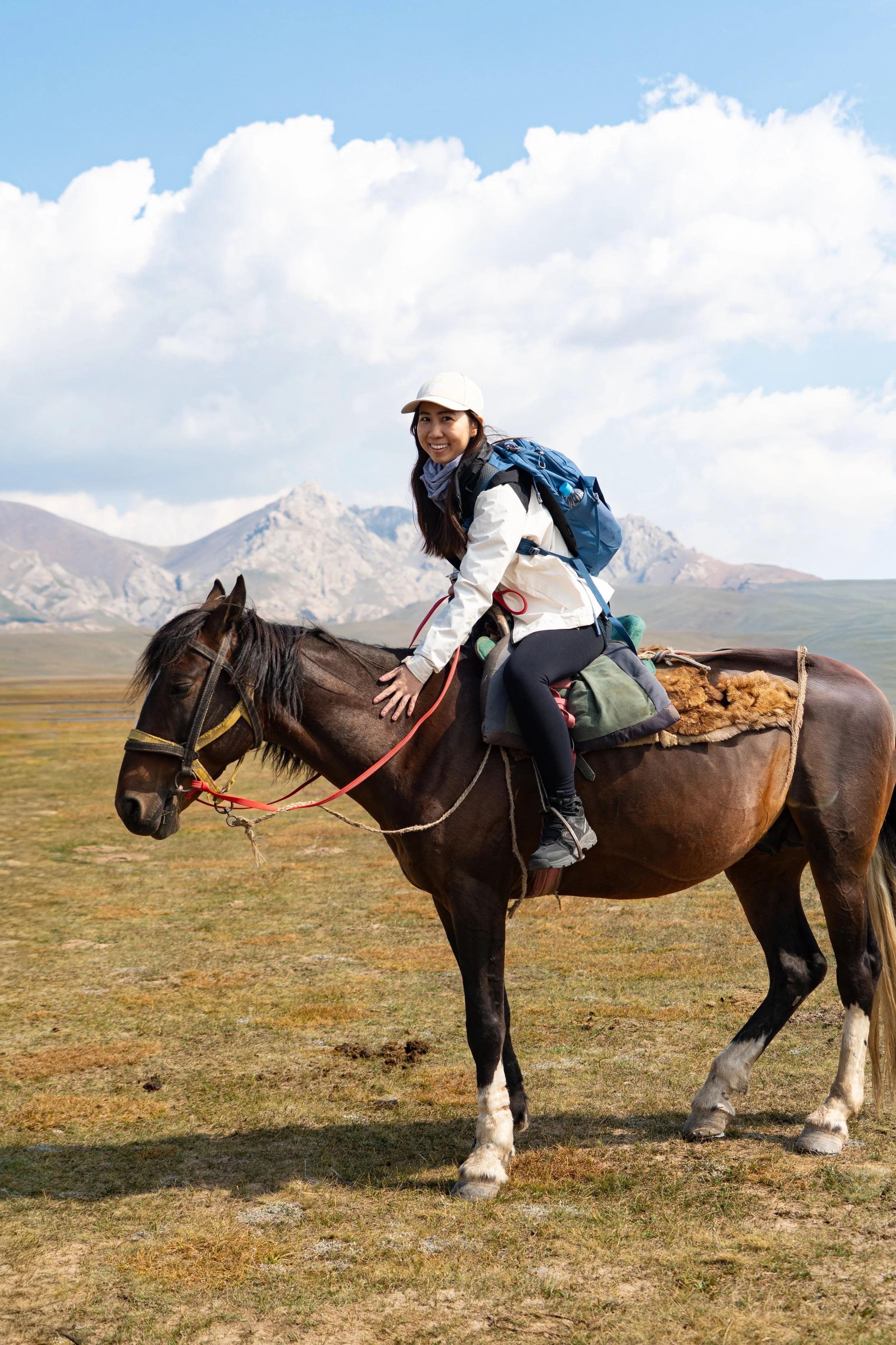 A woman riding a horse in a grassland with mountains and clouds in the background.
