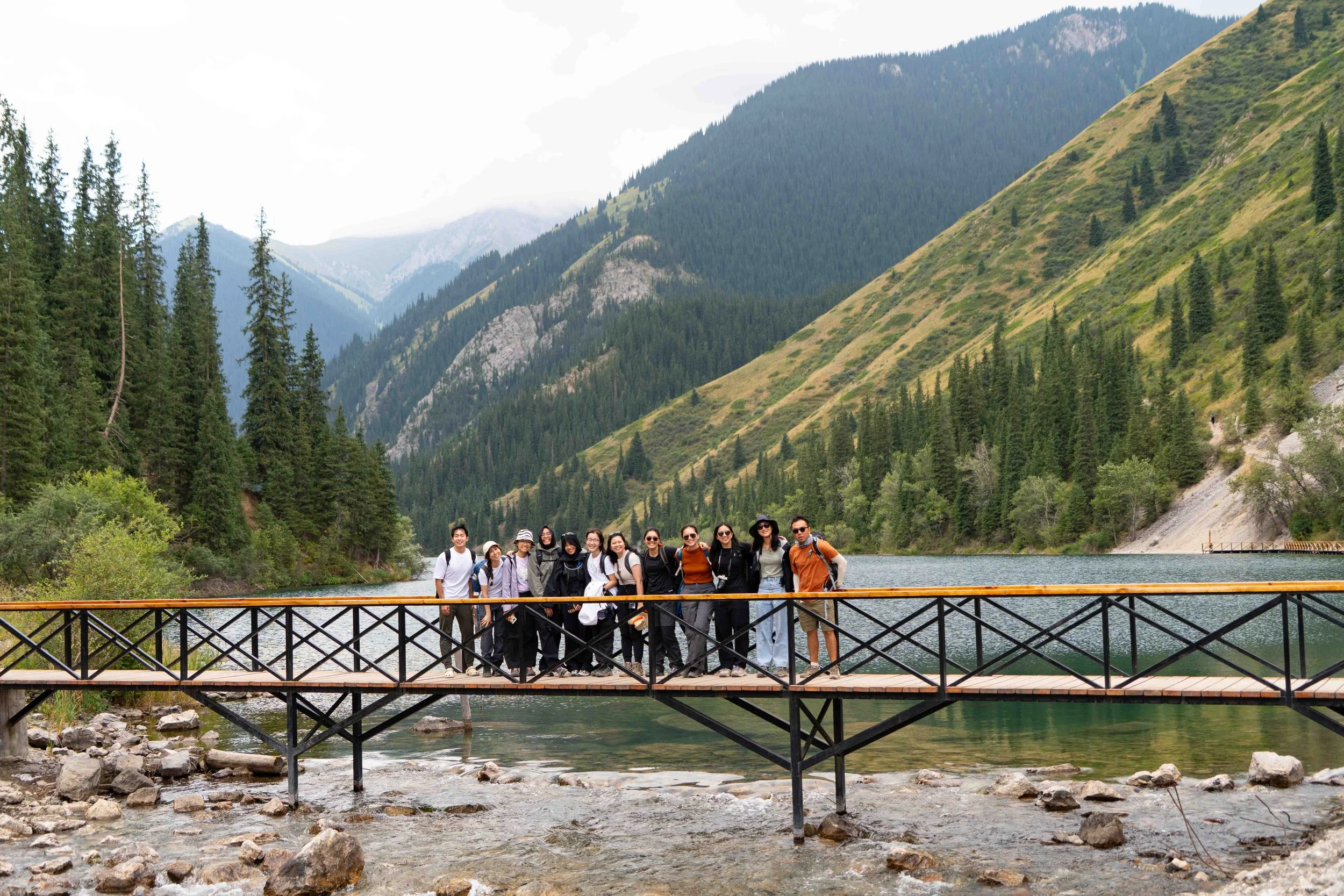 Kazakhstan, Kolsay lake A group of people standing on a wooden bridge over a river, surrounded by mountains and dense green pine forests.