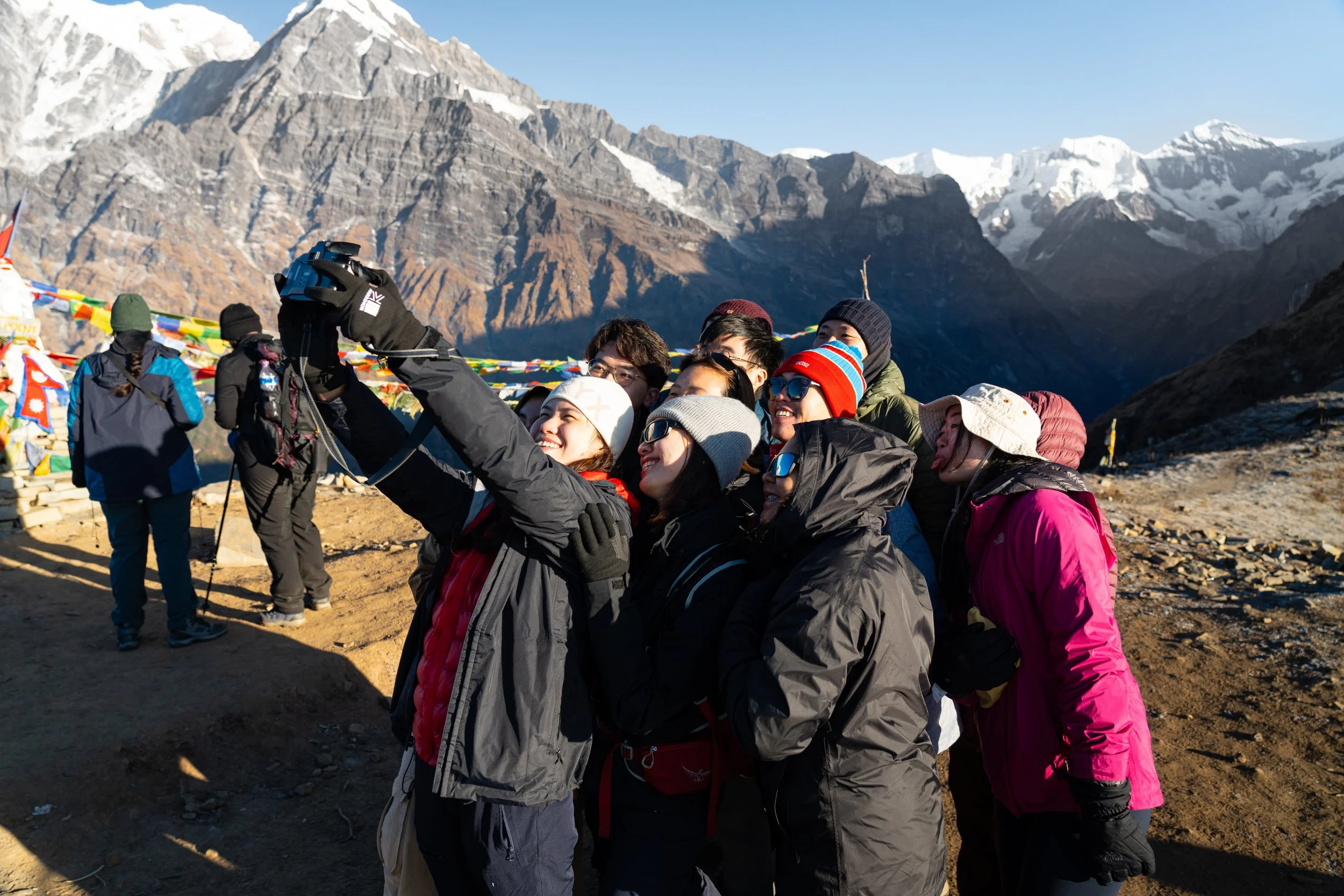 Group of people in winter clothing taking a selfie with mountains, snow, and prayer flags in the background.