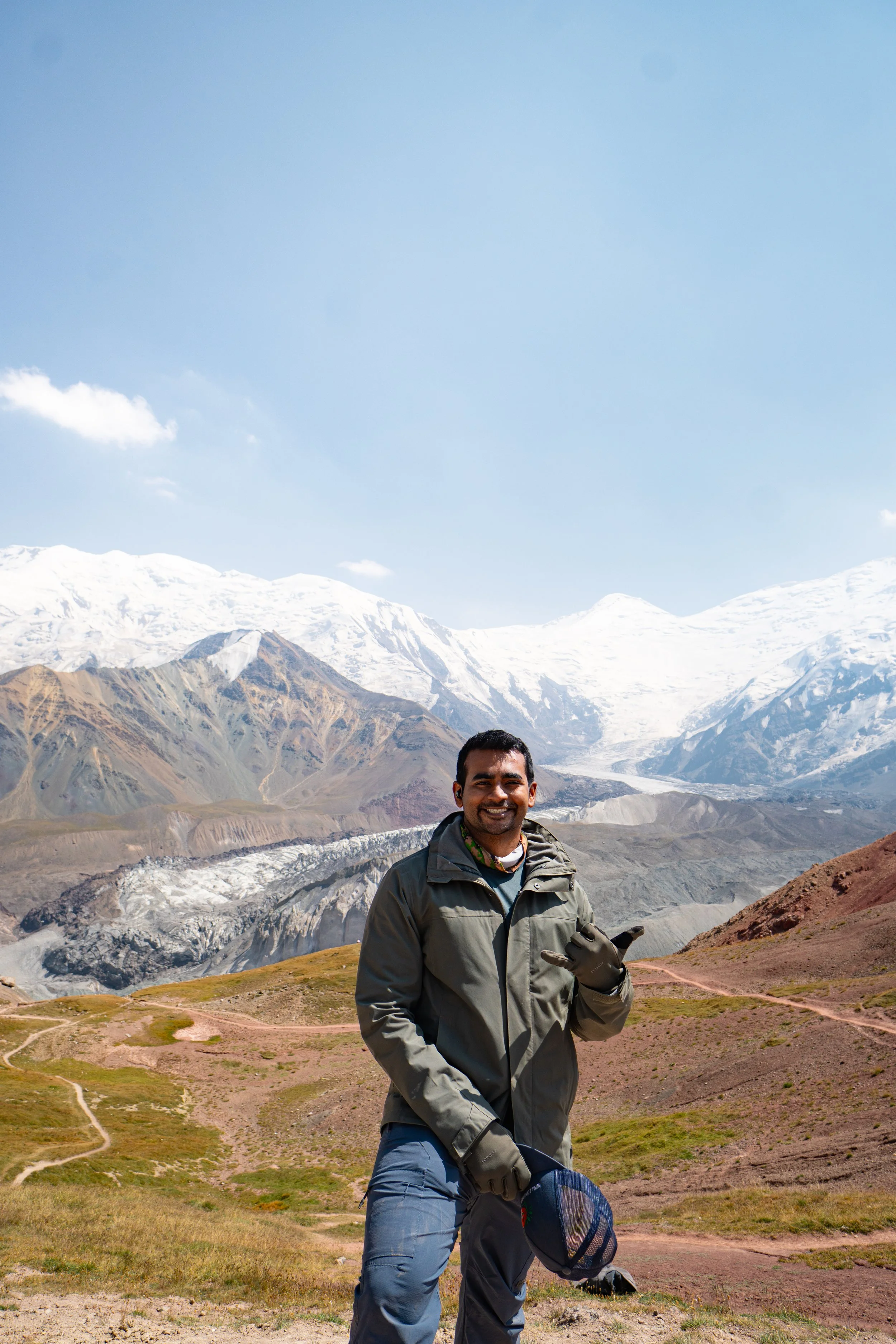 A man standing outdoors with mountains and glaciers in the background, smiling and holding a hat.