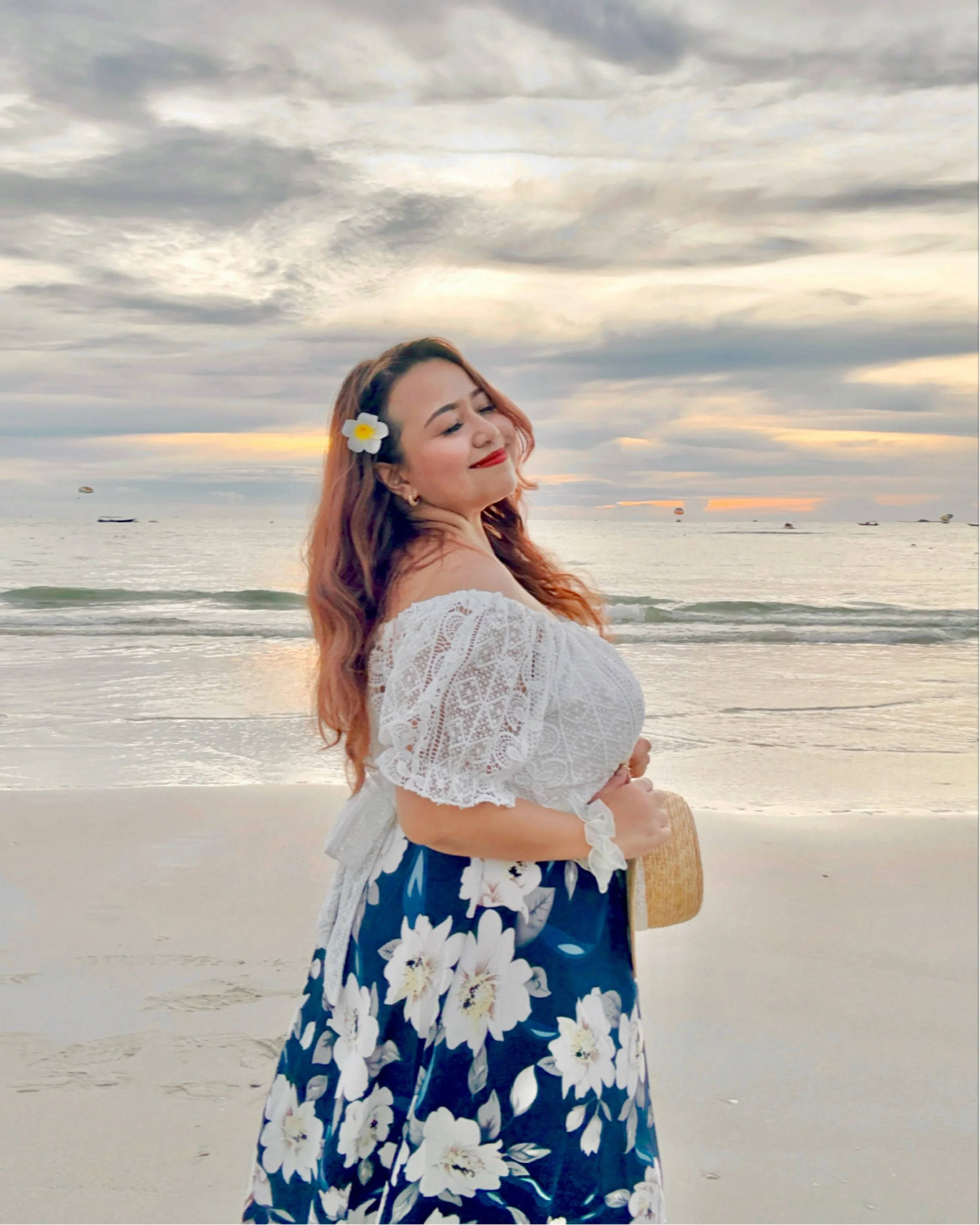 Woman with long wavy hair, wearing a white lace off-shoulder top and a floral skirt, standing on a beach at sunset with a flower in her hair.