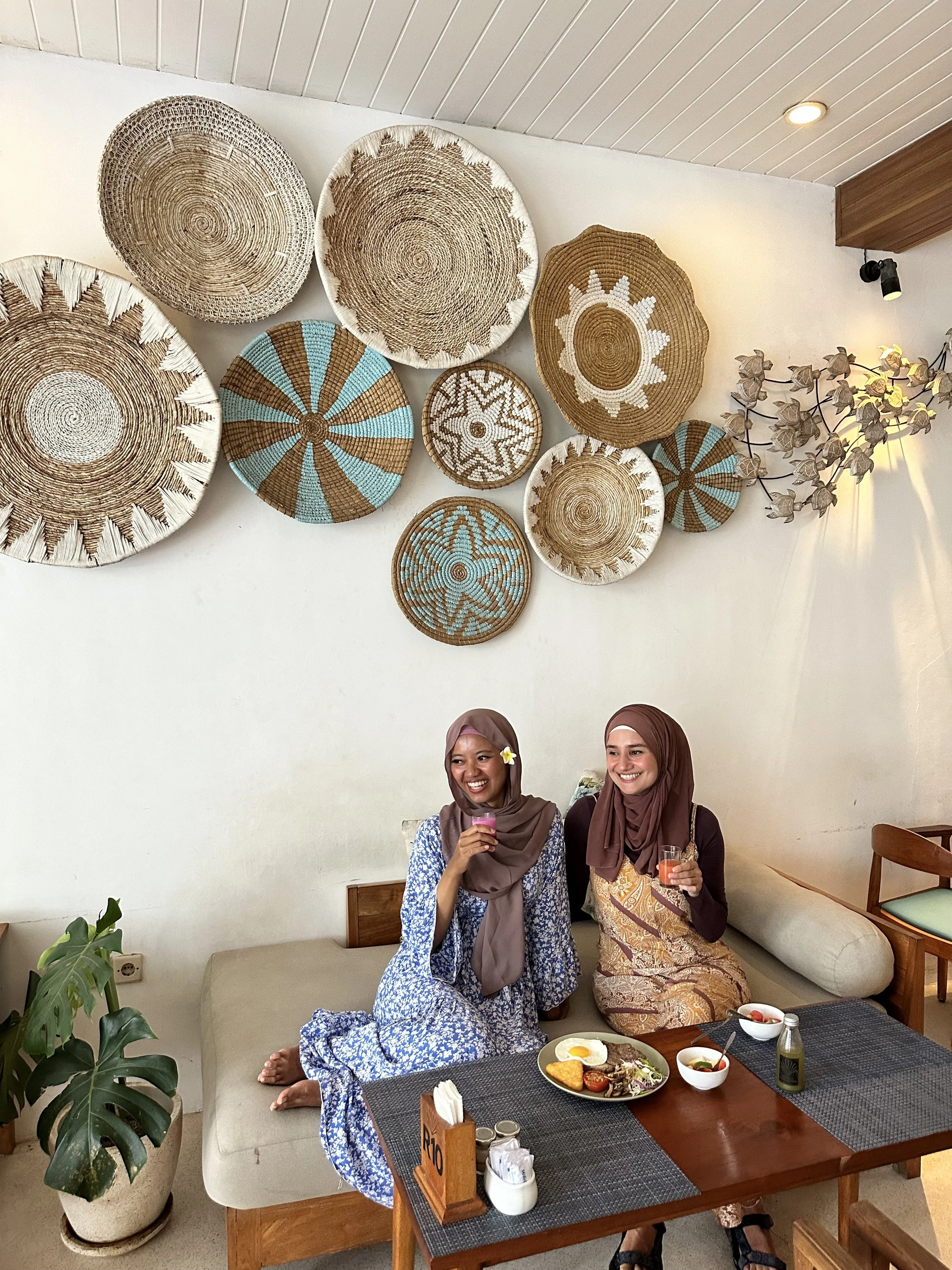 Two women wearing headscarves sitting on a beige sofa in a cafe, smiling and holding drinks, with a wooden table set with food and condiments in front of them, and decorative woven wall art and a potted plant in the background.