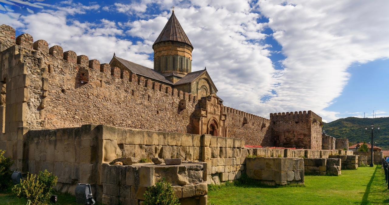 Ancient stone fortress walls with a church tower and a conical roof, green grass, and a partly cloudy sky.