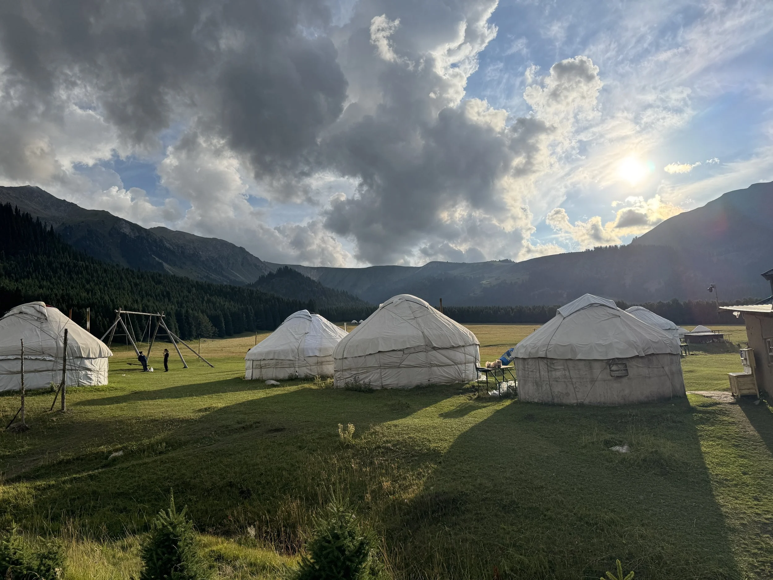 Kyrgyzstan  Green meadow with white yurts, mountains in the background, cloudy sky, and the sun partially visible.
