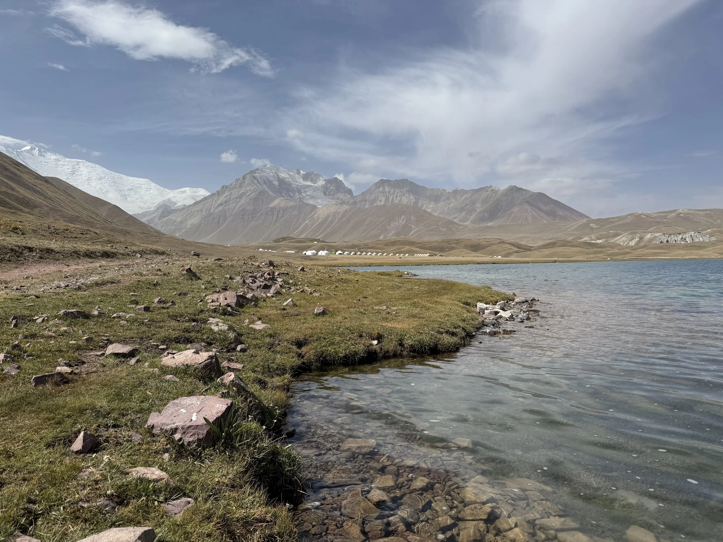 Scenic view of a lake with clear water and rocky shoreline, surrounded by grassy land and mountains in the background, partly snow-capped, under a partly cloudy sky.