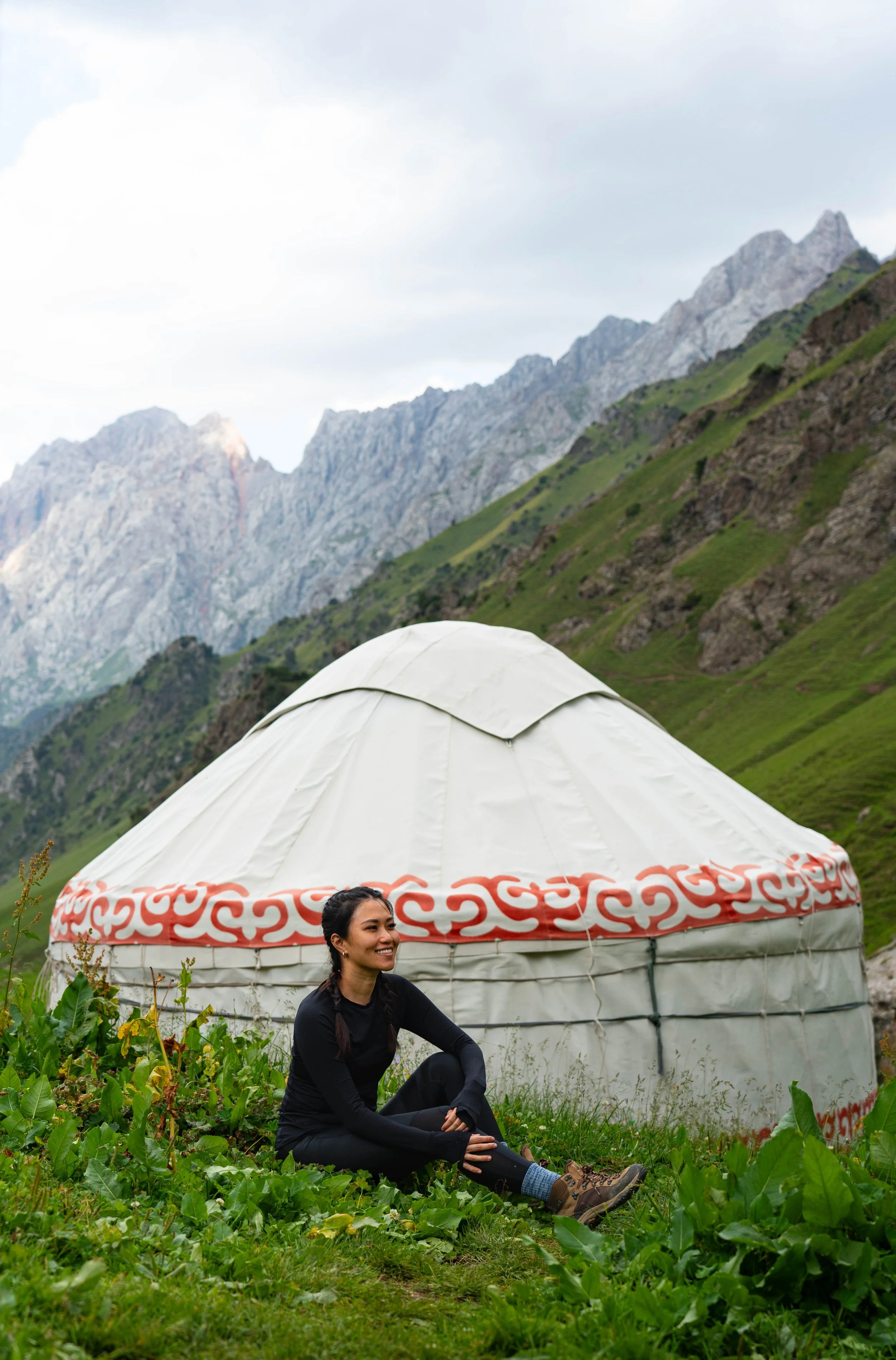 A woman sitting on grass with a smile in front of a traditional yurt, surrounded by green plants, with mountains in the background.