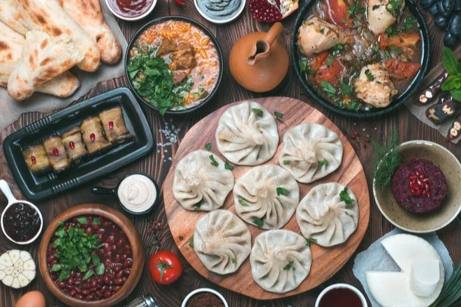 Georgia Cuisine. Assorted dishes including dumplings on a wooden board, a bowl of red beans, a plate with stuffed grape leaves, a bowl of tomato-based stew, a bowl of soup, various sauces, bread, and garnishes on a dark wooden table.