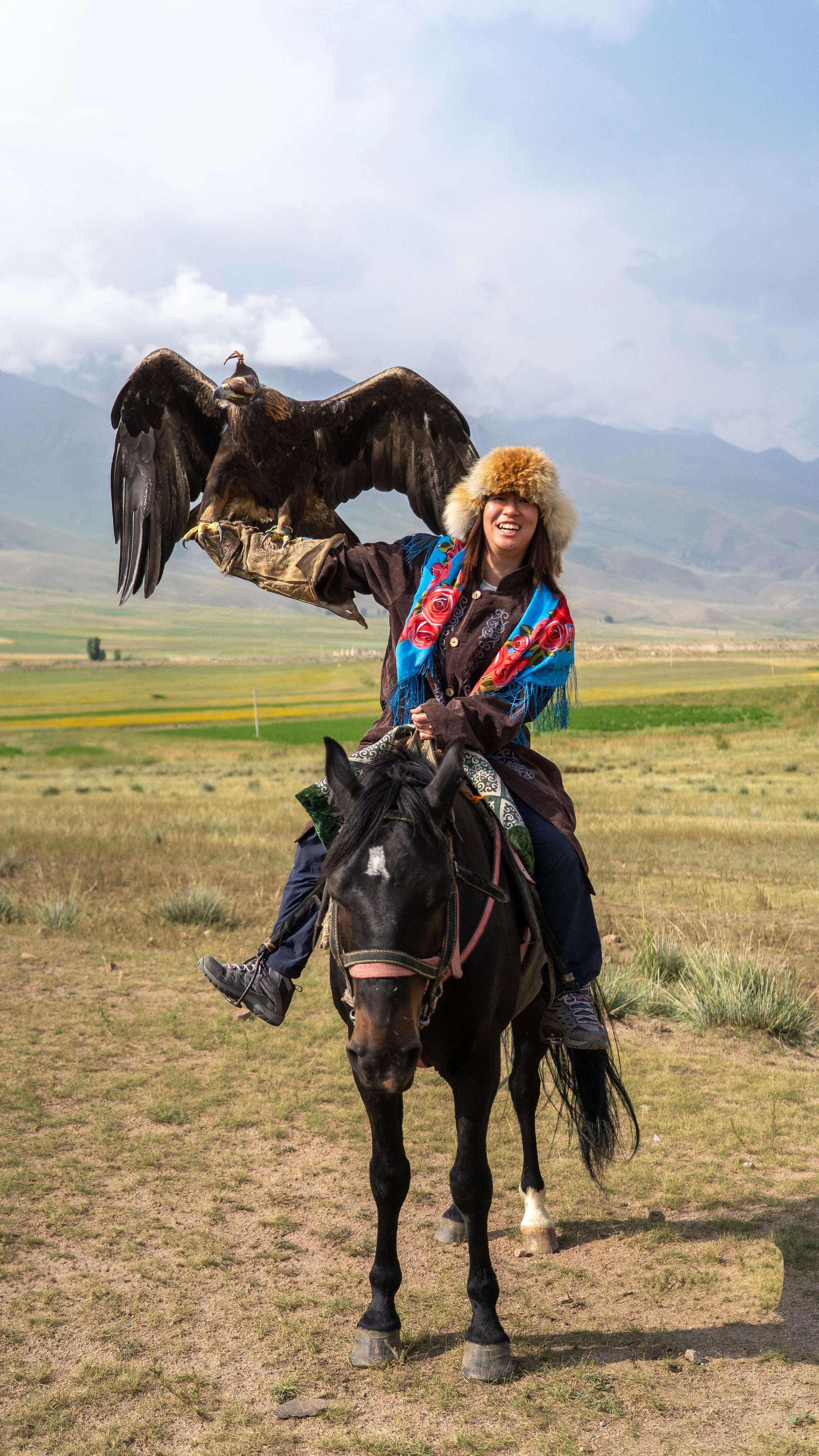 Kazakhstan. A woman wearing a fur hat and colorful scarf riding a black horse in a grassy plain with mountains in the background, holding a large bird of prey on her arm.
